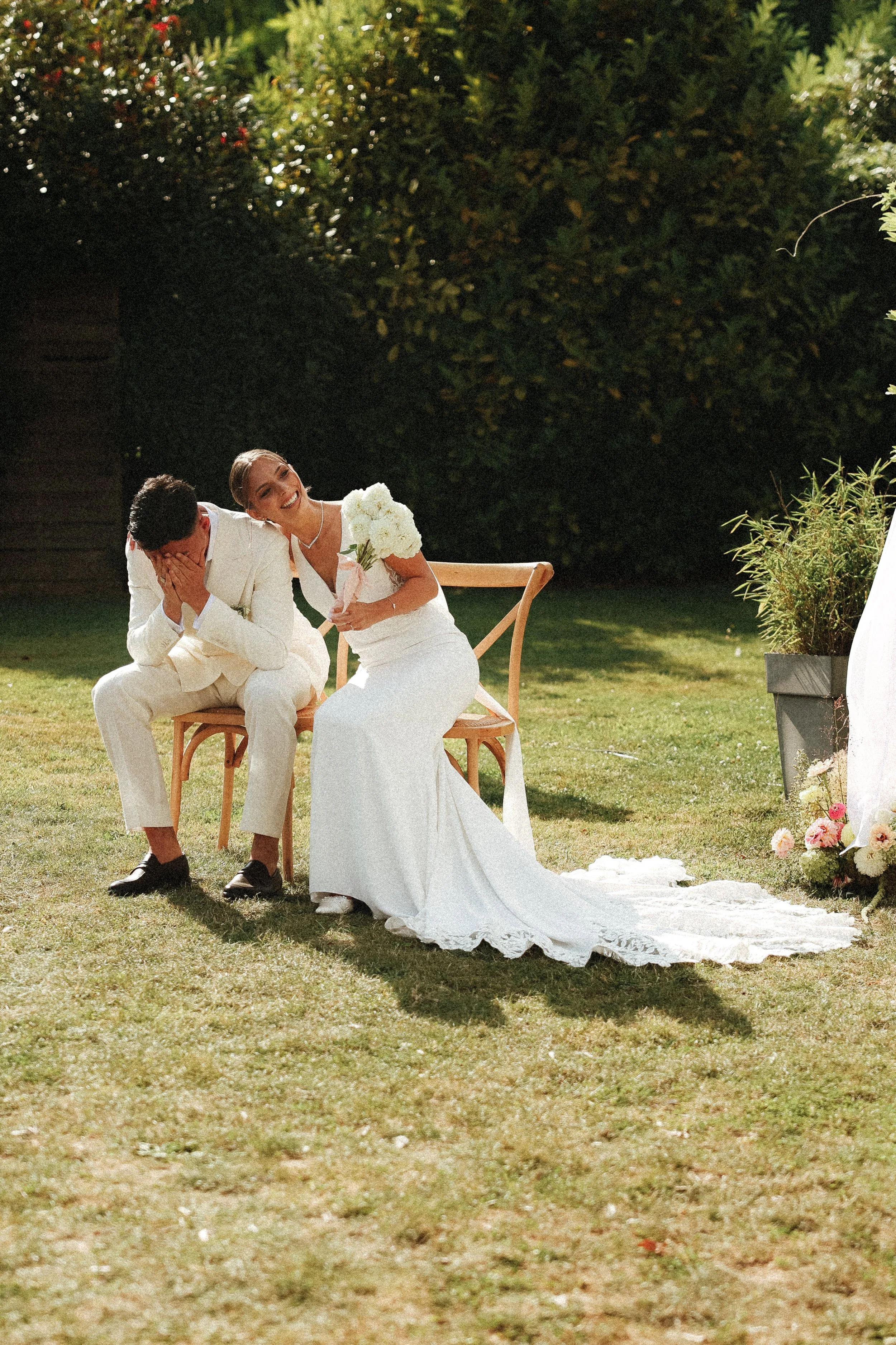 Un couple en vêtements de mariage, assis sur une chaise dans un jardin, l'épouse rit avec un bouquet de fleurs dans la main, l'époux couvre son visage de ses mains, ambiance lumineuse en plein air.