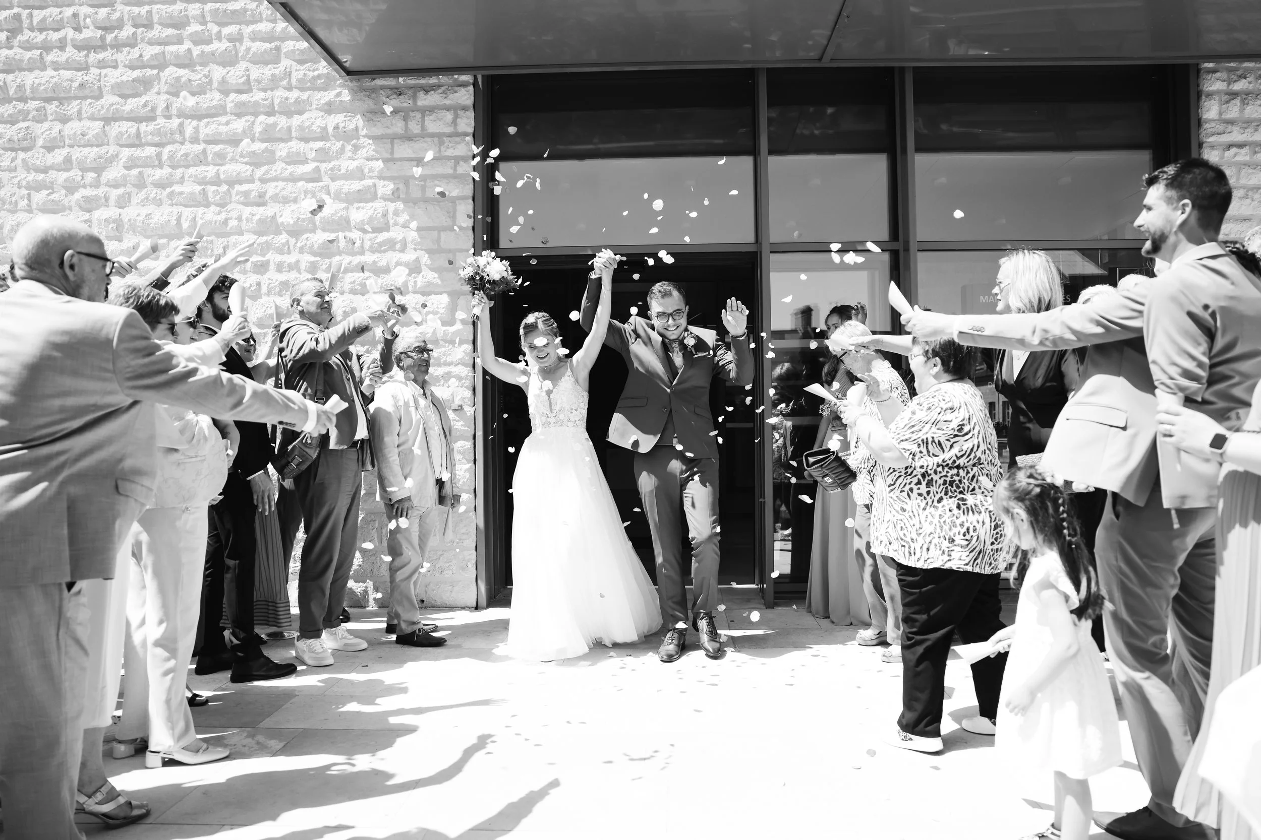 Un groupe de personnes, incluant un couple marié, fête à l'extérieur devant un bâtiment en pierre, alors que des invités lancent des pétales de fleurs en l'air pour célébrer leur mariage.
