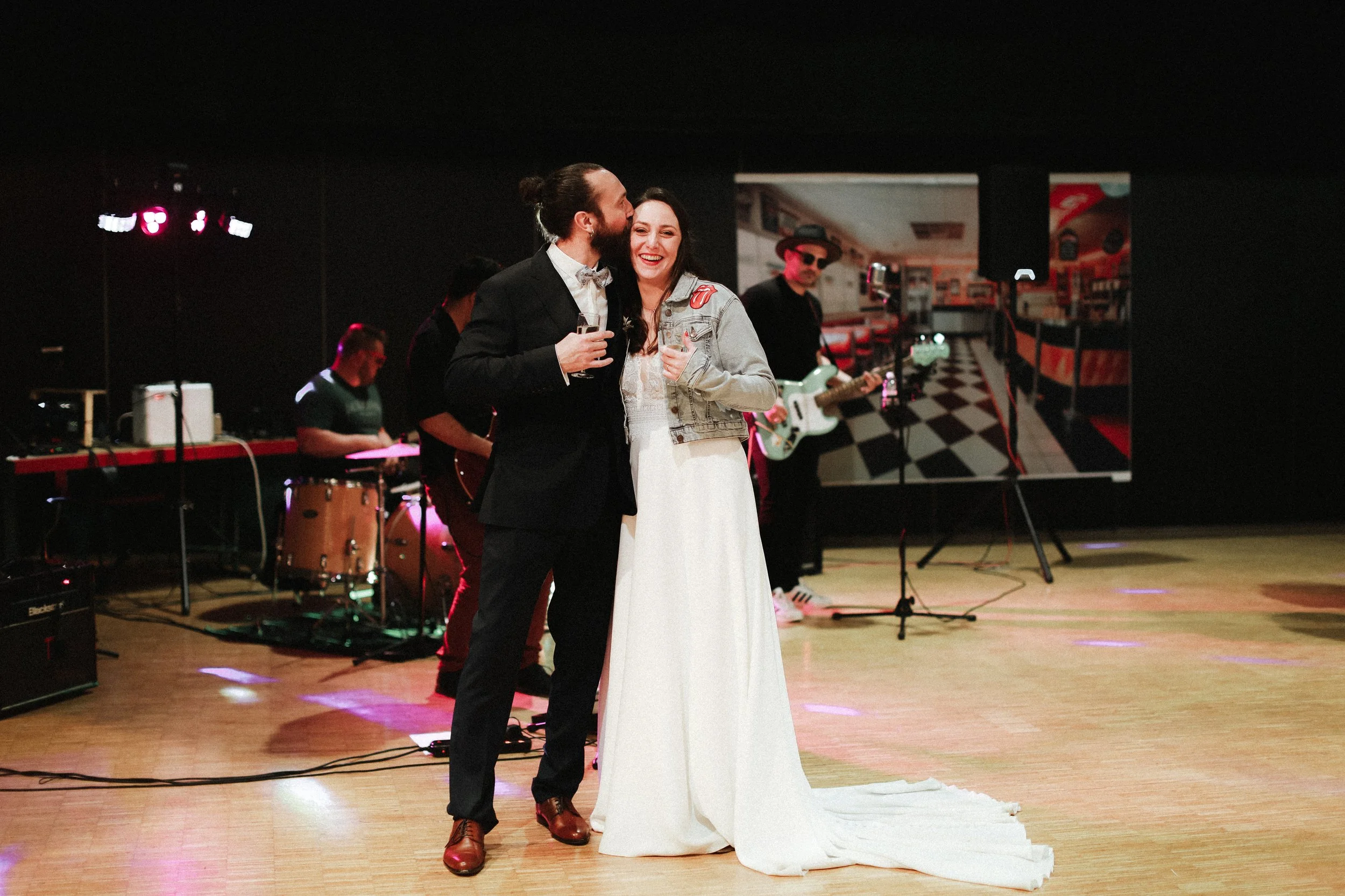 Un couple de mariés, un homme et une femme, sourient et se tiennent dans une salle de réception avec un groupe de musiciens en arrière-plan, pendant leur mariage.