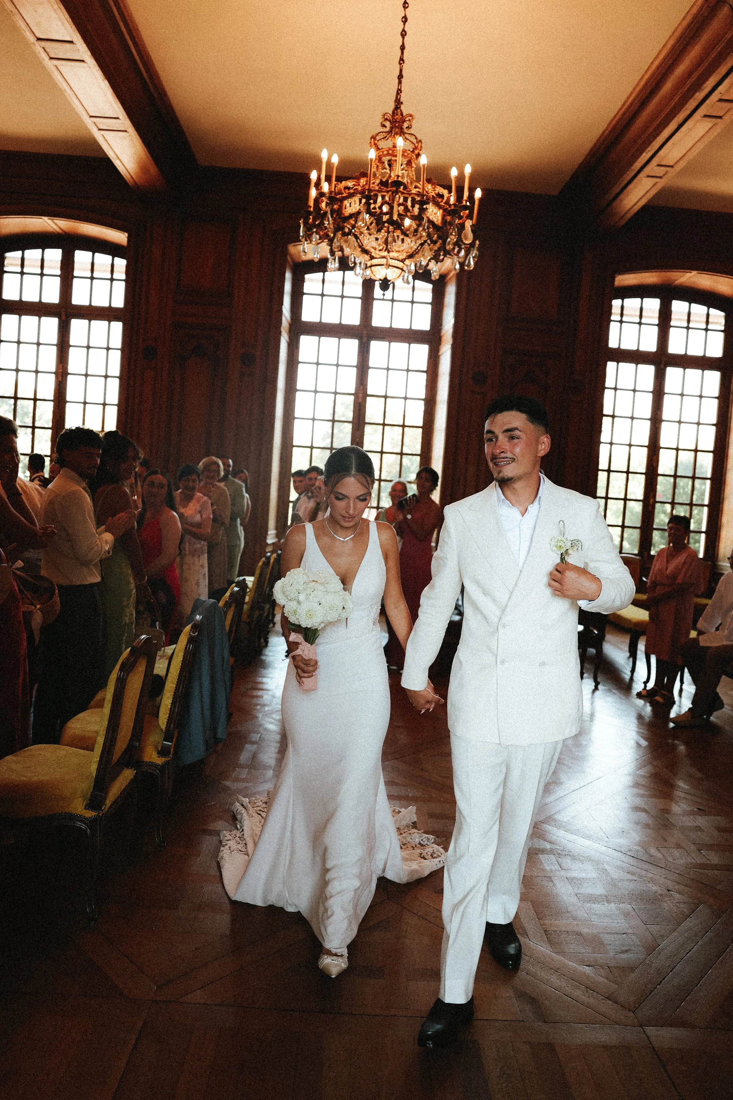 Un couple en tenue de mariage marche main dans la main dans une salle avec de grandes fenêtres et un chandelier en cristal suspendu au plafond, entourés de leurs invités.
