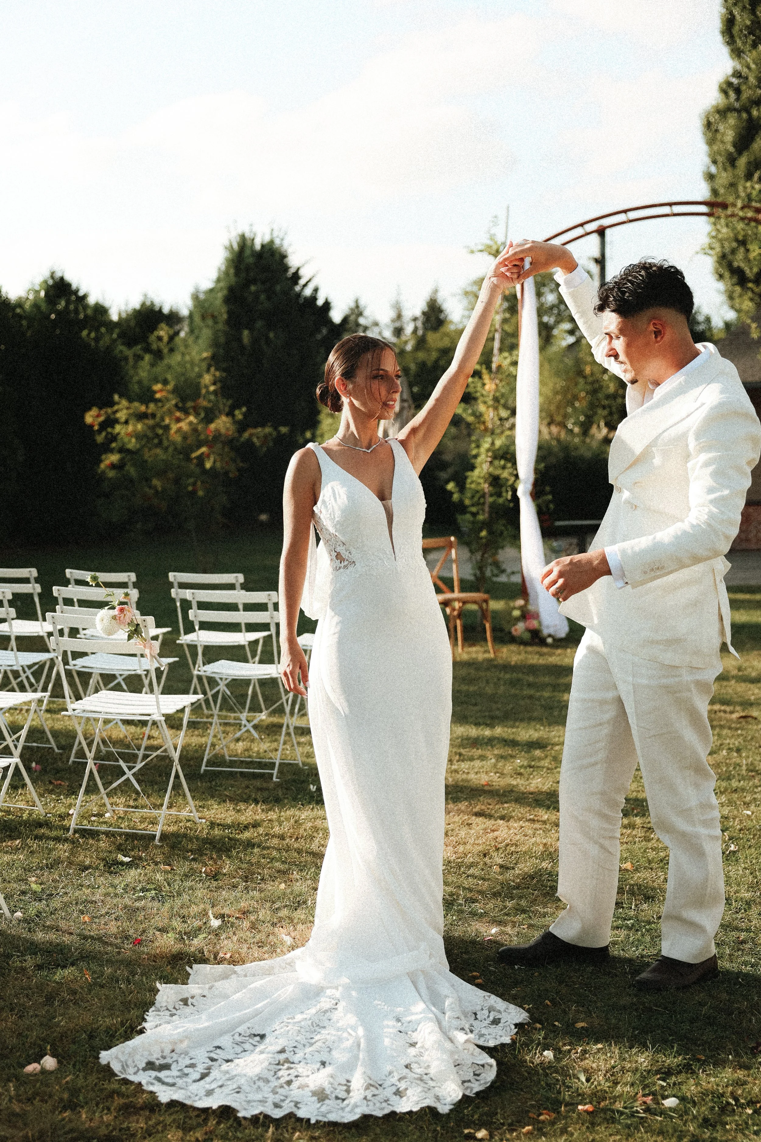 Un couple en vêtements de mariage danse lors d'une cérémonie en plein air, avec des chaises blanches décorées de fleurs derrière eux.
