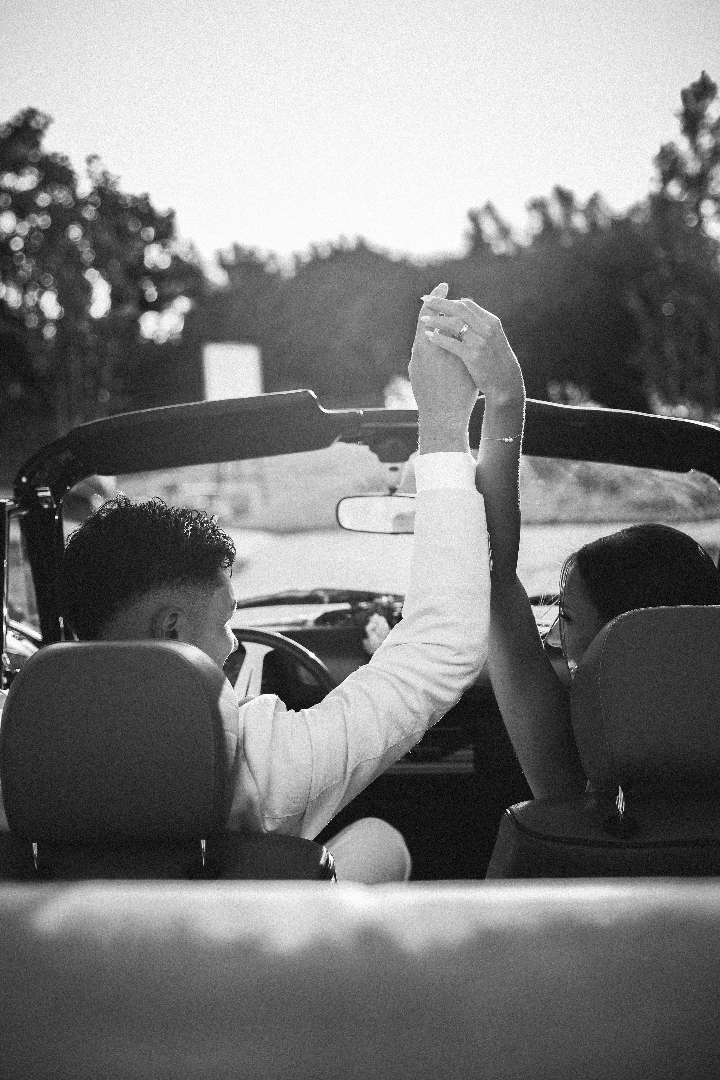 Un couple assis dans une voiture, se tenant la main levée. Le fond est en extérieur avec des arbres, et la photo est en noir et blanc.