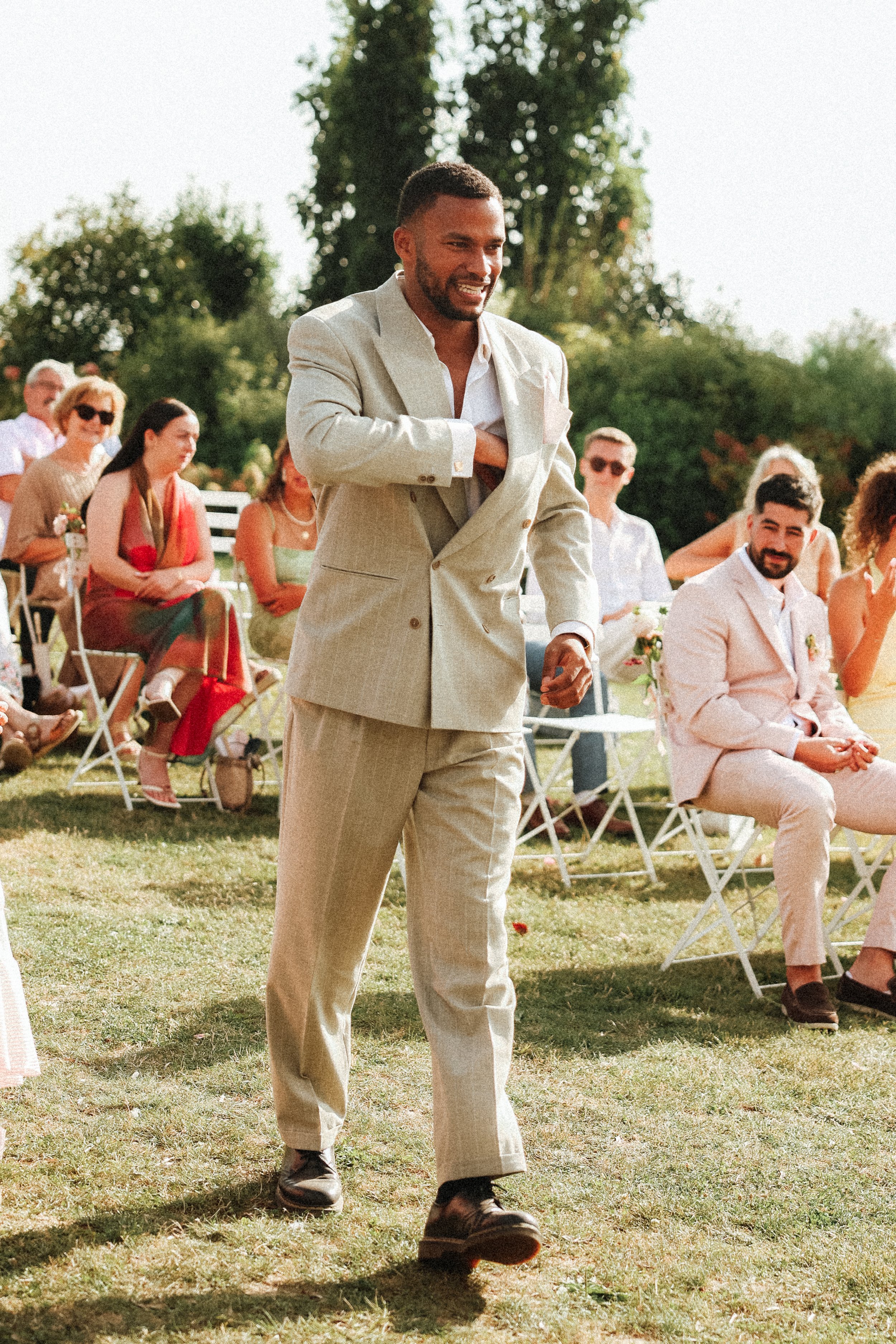 Homme souriant en costume beige lors d'un mariage en plein air, entouré de personnes assises et de la nature.