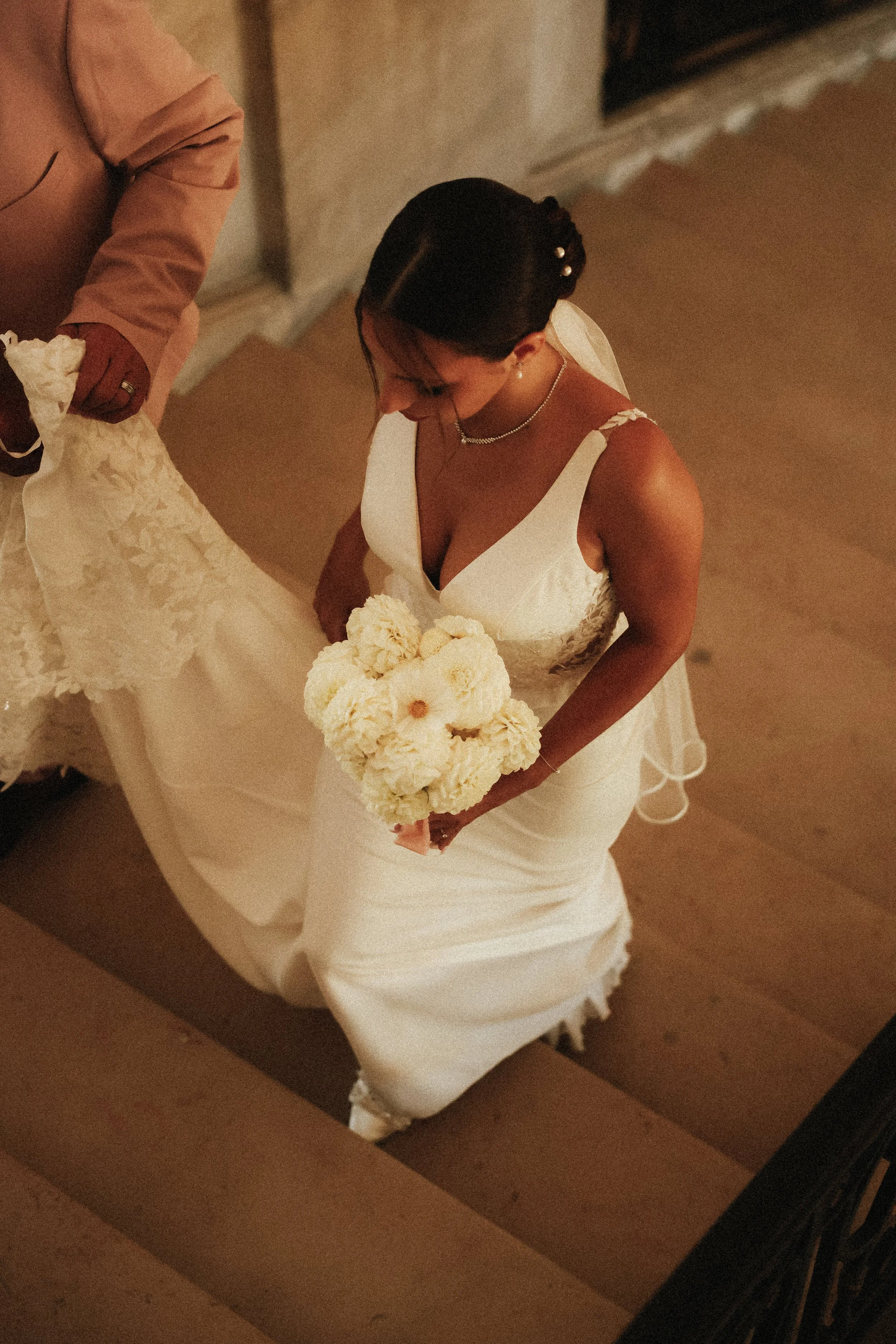 Une mariée en robe blanche tenant un bouquet de fleurs blanches, vue de haut dans une salle avec un escalier en bois.
