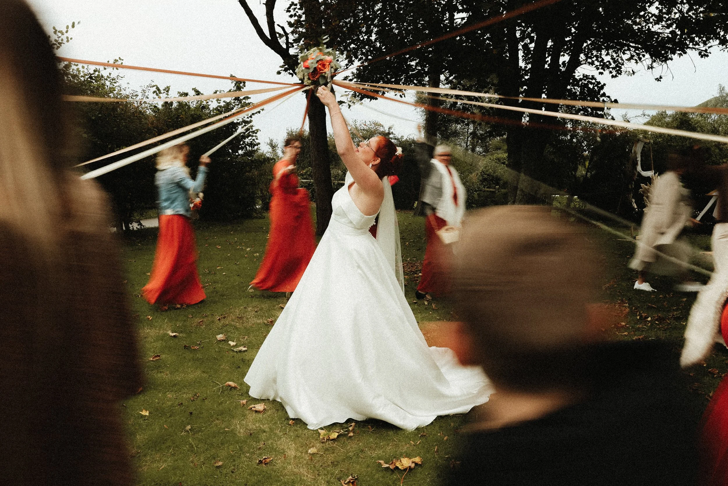 Une mariée en robe blanche accroche un bouquet de fleurs à un poteau lors d'une fête en extérieur, entourée de plusieurs personnes en mouvement, dans un cadre naturel avec des arbres.