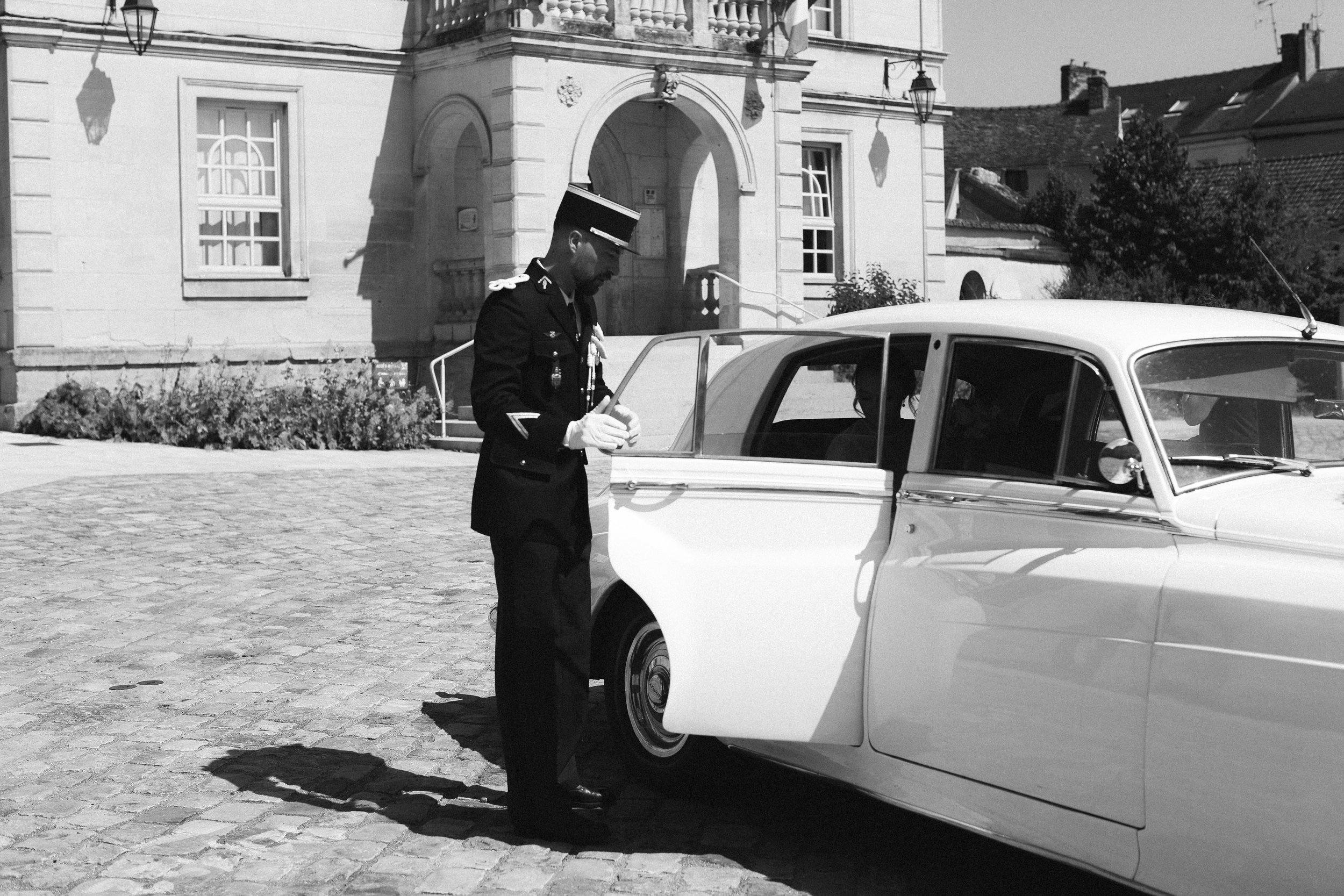 Un officier en uniforme de police écrivant quelque chose près d'une voiture ancienne blanche devant un bâtiment en pierre.