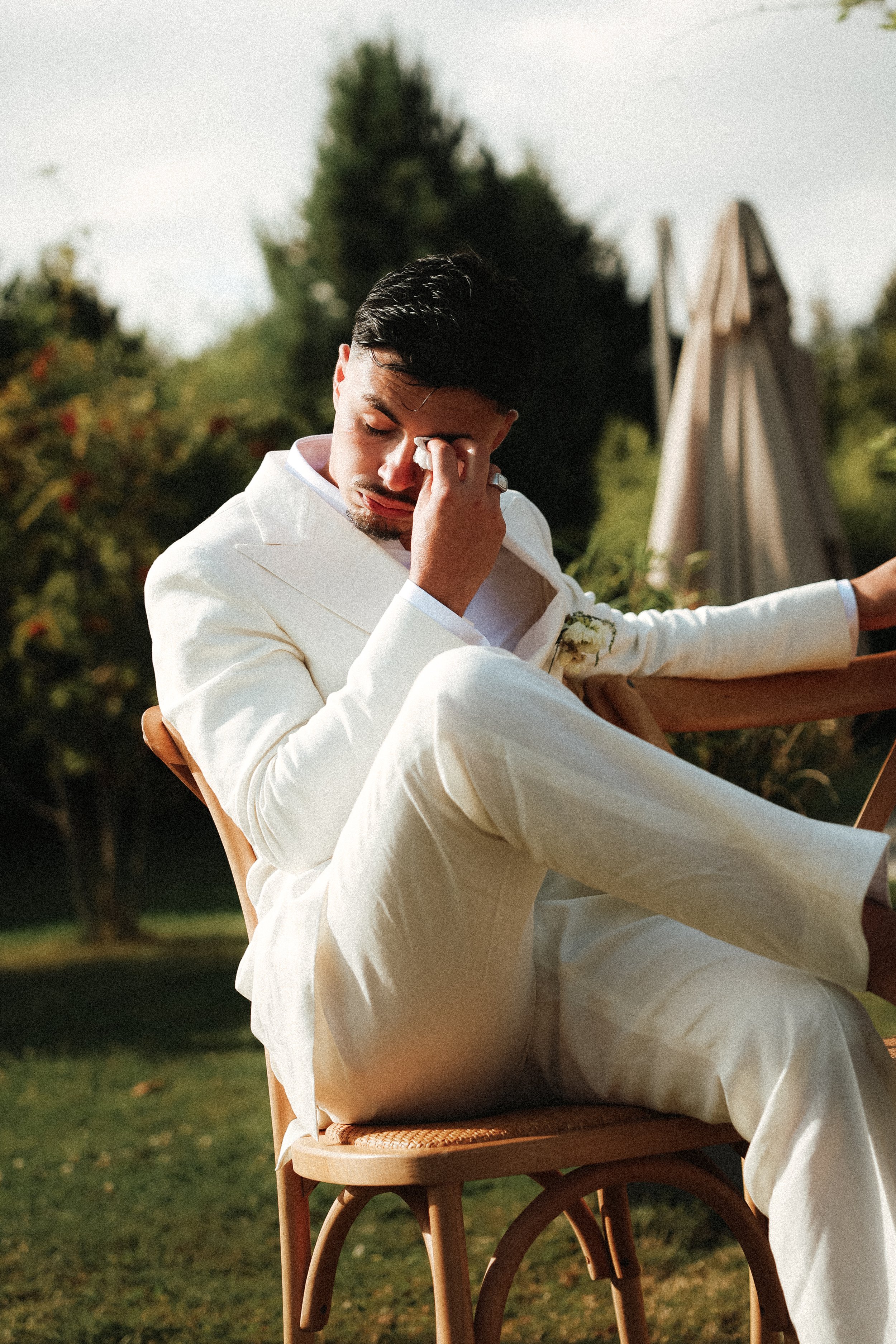 Un homme en costume blanc assis sur une chaise en bois dans un jardin, l'air bouleversé, se frottant les yeux.