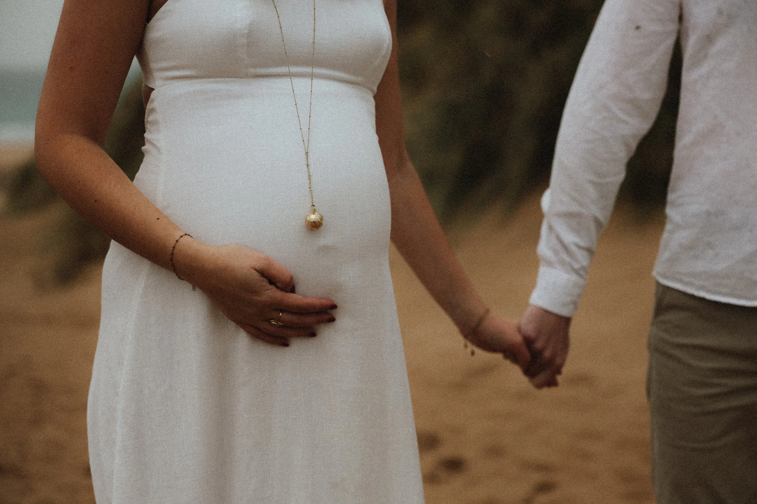 Une femme enceinte en robe blanche tient la main d'un homme lors d'une séance photo sur une plage.