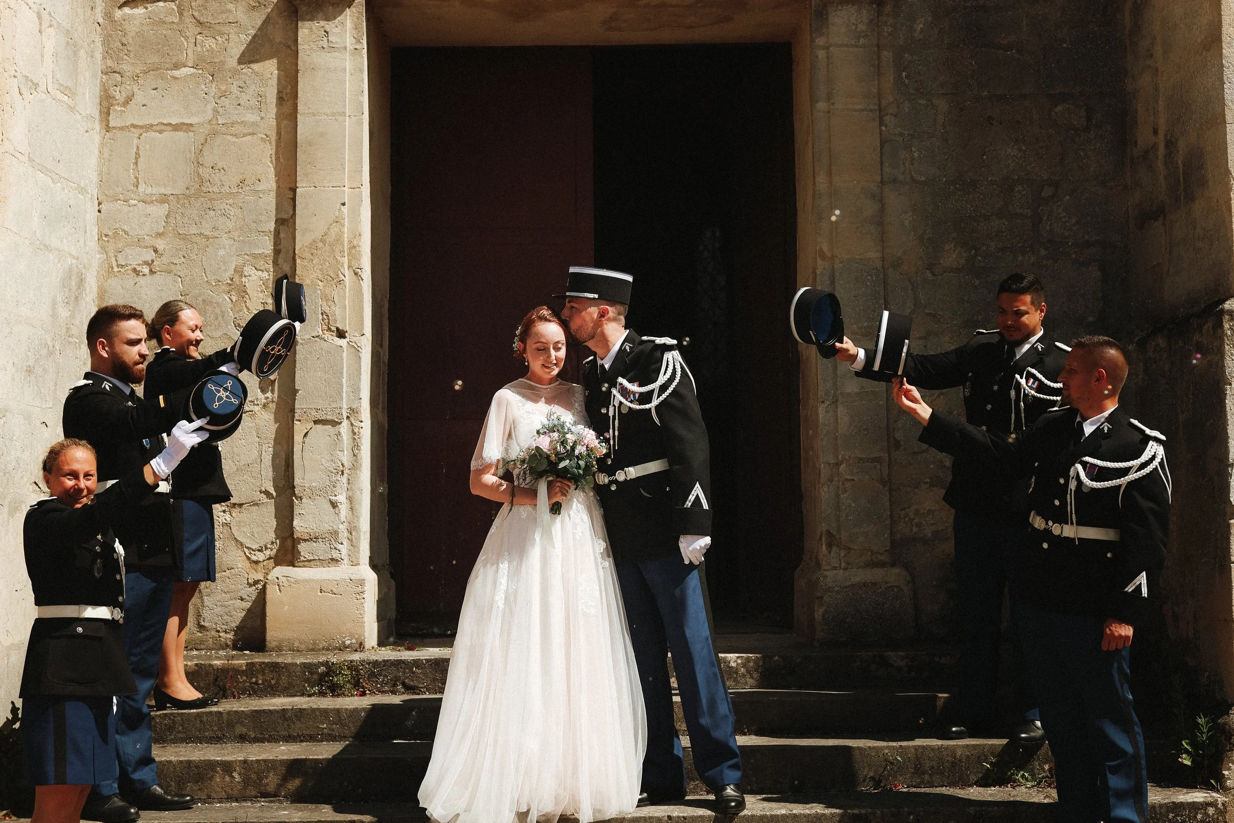 Un couple de mariés, un homme en uniforme militaire, partage un moment intime en dehors d'une vieille église, entourés de personnes en uniforme qui ont levé leurs chapeaux en signe de respect ou de célébration.