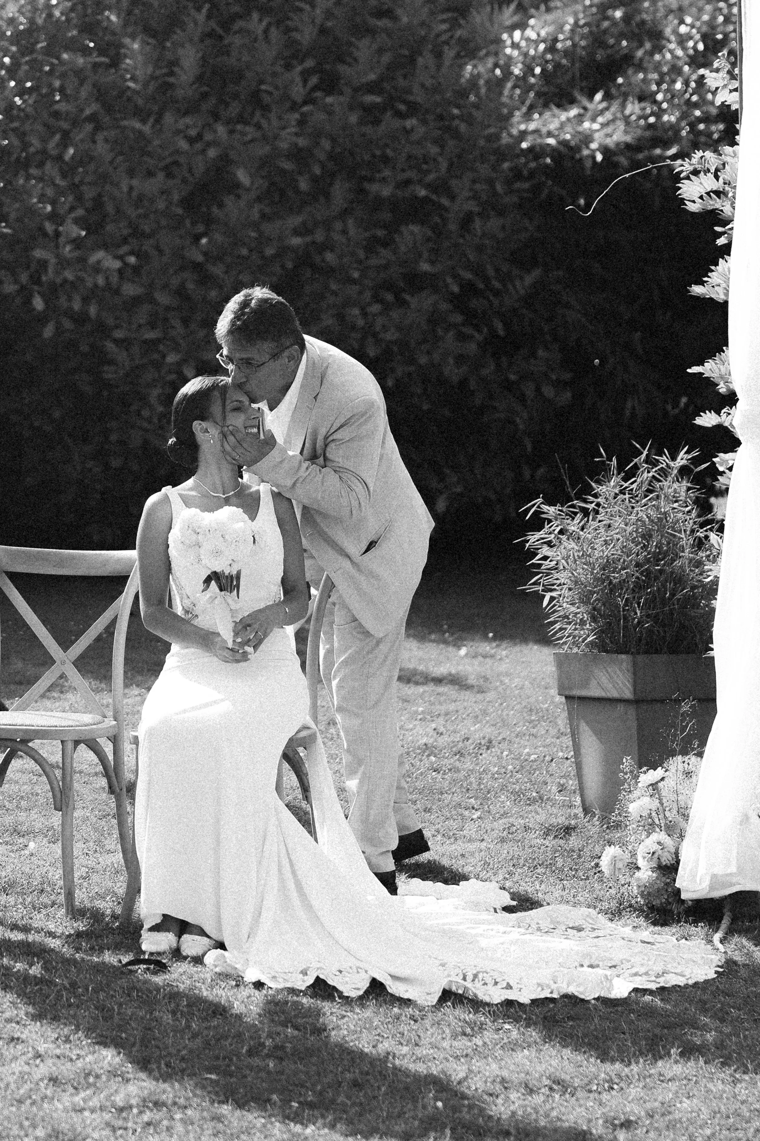 Un couple à un mariage en extérieur, où l'homme embrasse la femme qui tient un bouquet de fleurs, lors d'une cérémonie nuptiale. La scène est en noir et blanc.