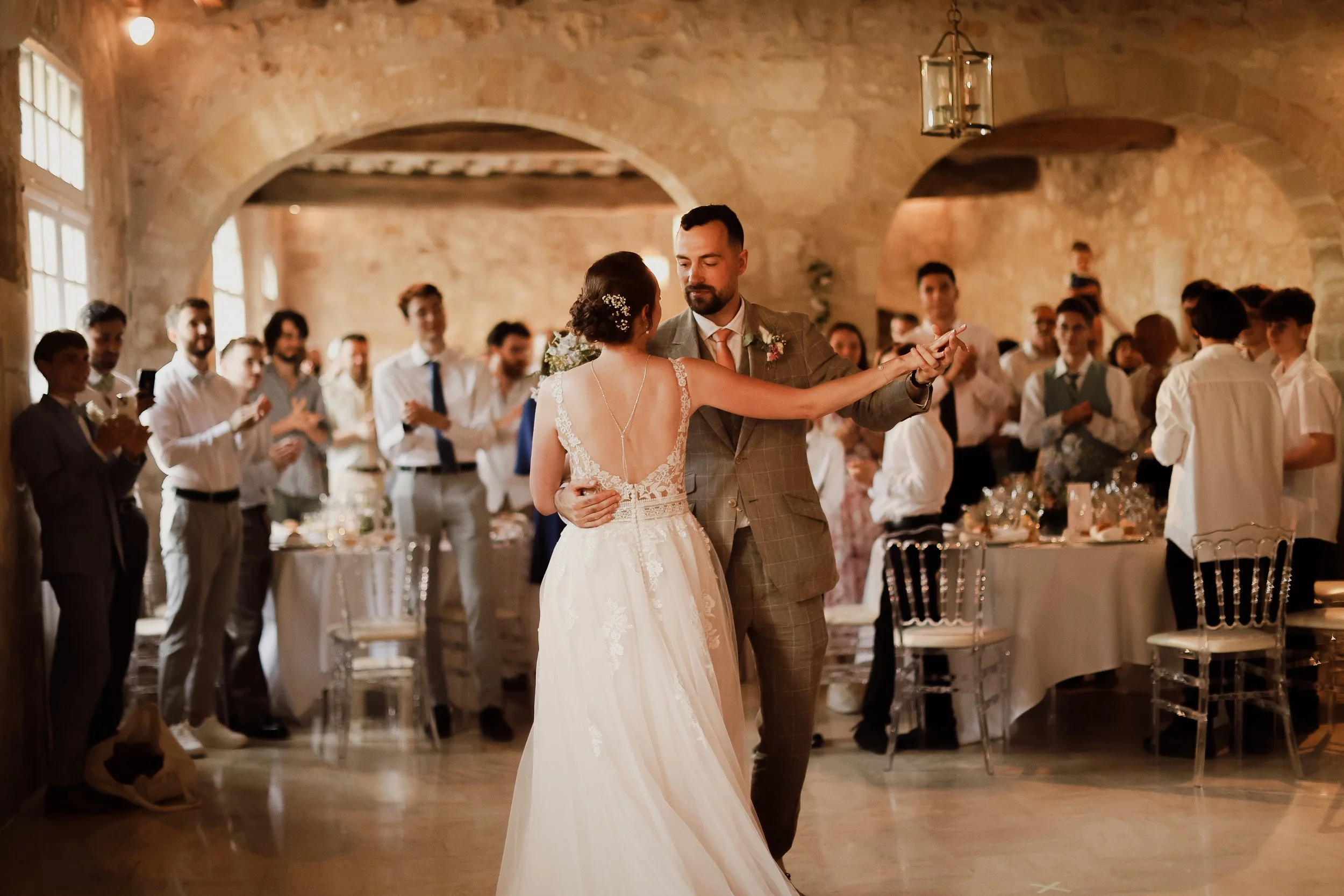 Un couple danse lors de leur mariage dans une salle en pierre, entouré d'invités qui les regardent et applaudissent.