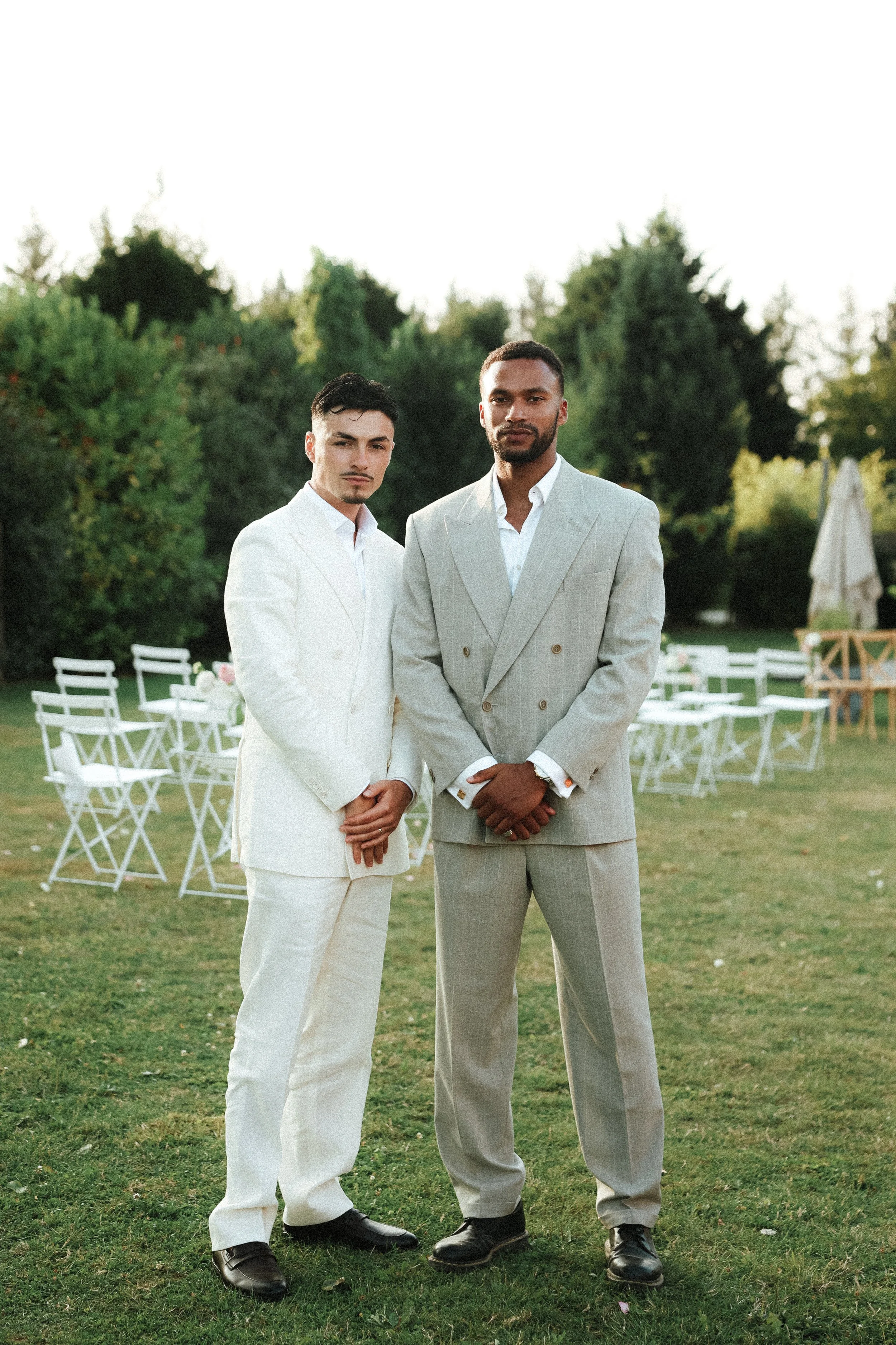 Deux hommes en costumes formels, devant des chaises blanches en extérieur, avec des arbres en fond.