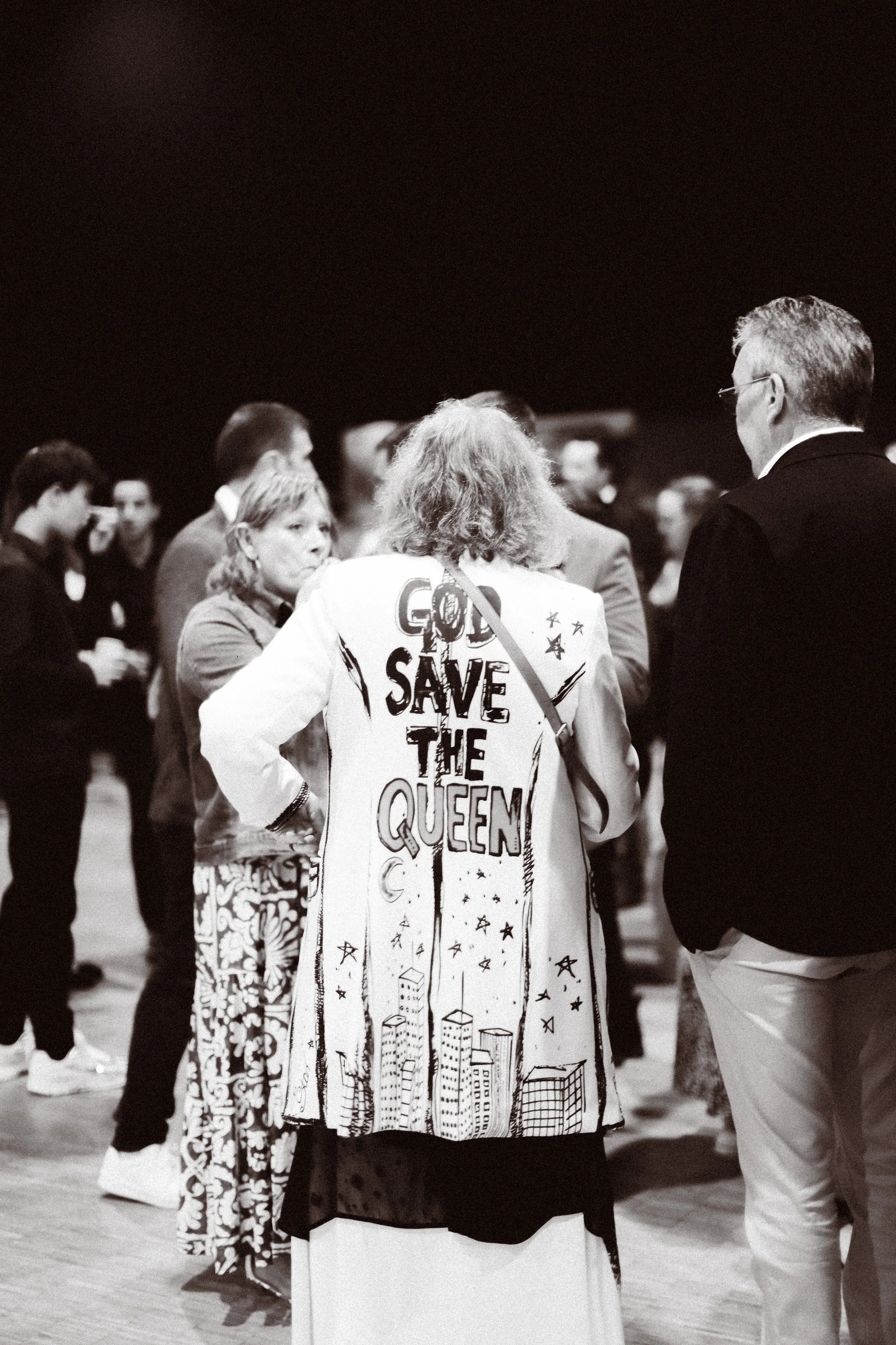 Personne portant une veste avec l'inscription "God Save The Queen", entourée de plusieurs personnes lors d'un événement.