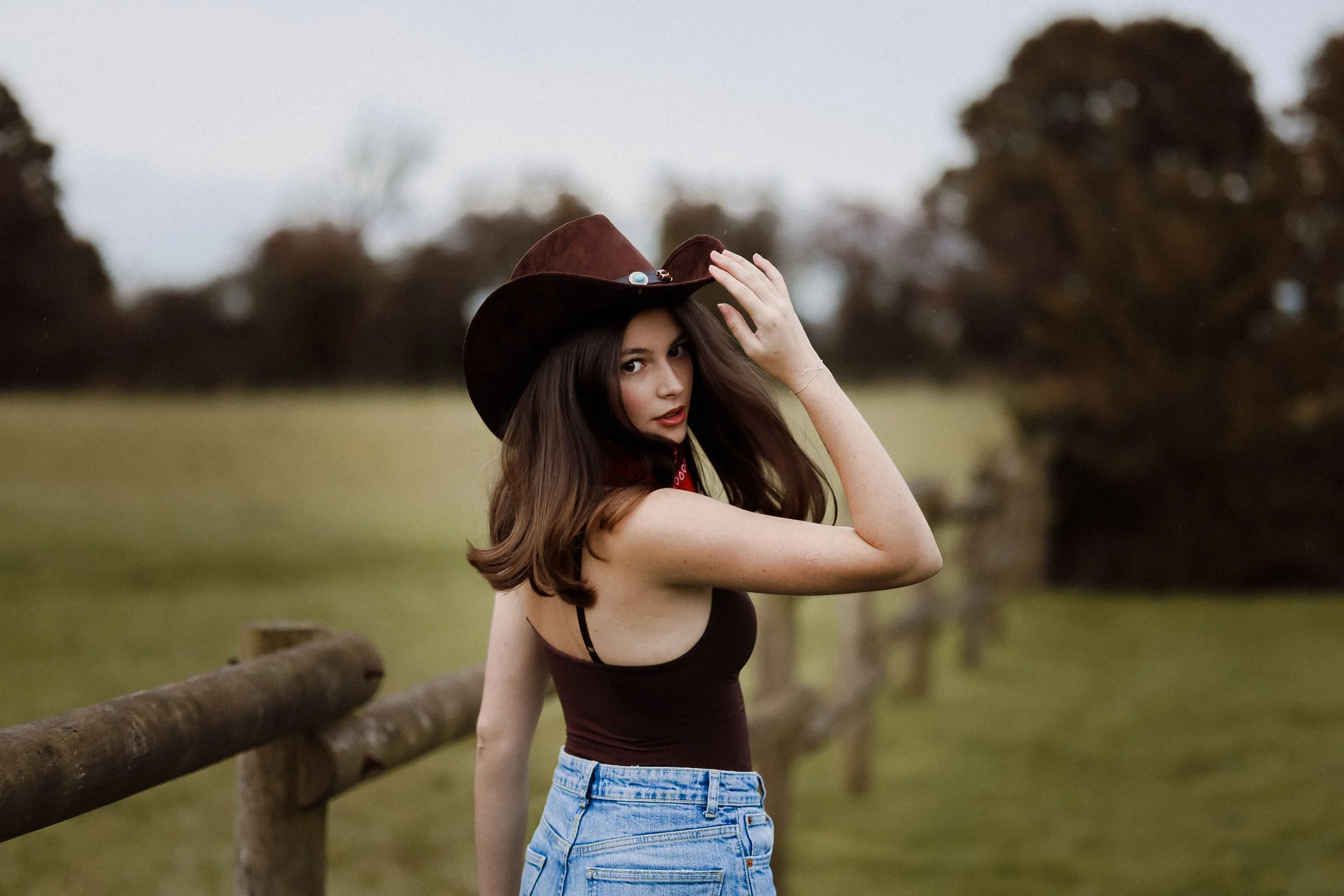 Jeune femme portant un chapeau de cowboy dans un champ avec une clôture en bois, au coucher du soleil. Séance portrait. Photographe Normandie. Séance portait.