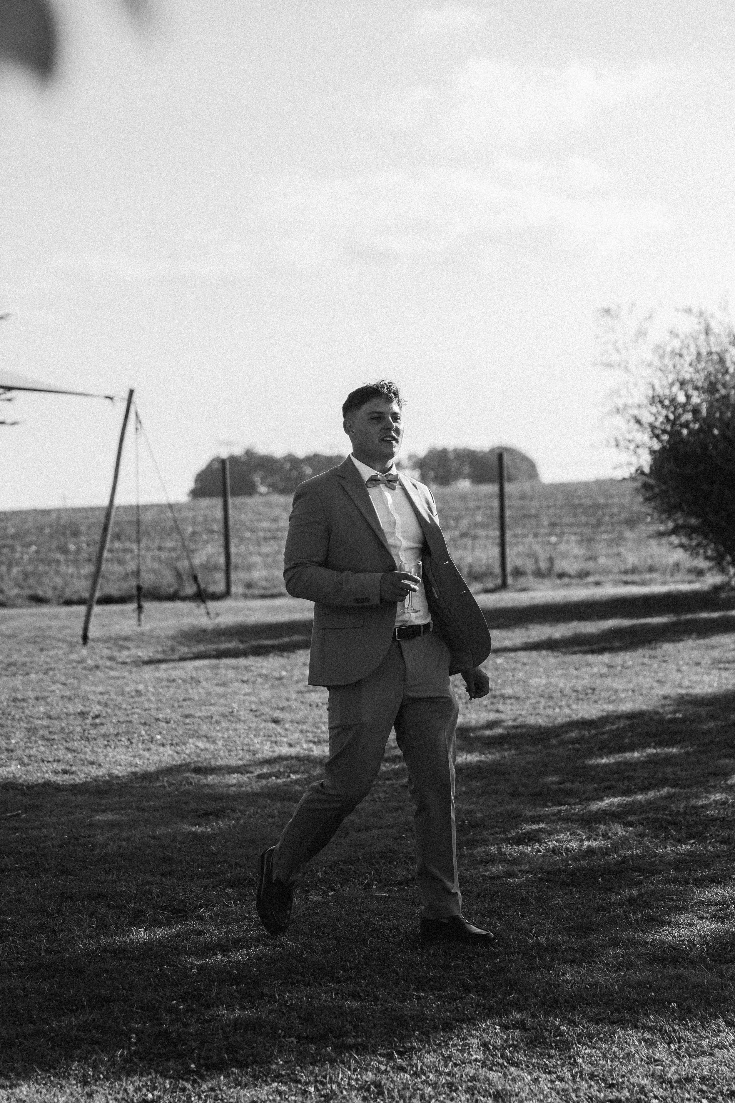 Un homme habillé en costume et nœud papillon marche dans un jardin, en noir et blanc.