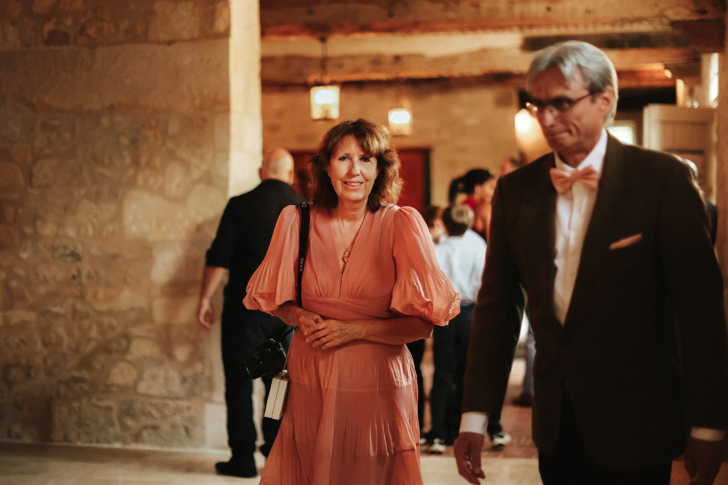 Une femme en robe rose pâle et un homme en smoking noir avec un nœud papillon dans une salle de réception avec des murs en pierre et des lumières suspendues, entourés d'autres personnes.