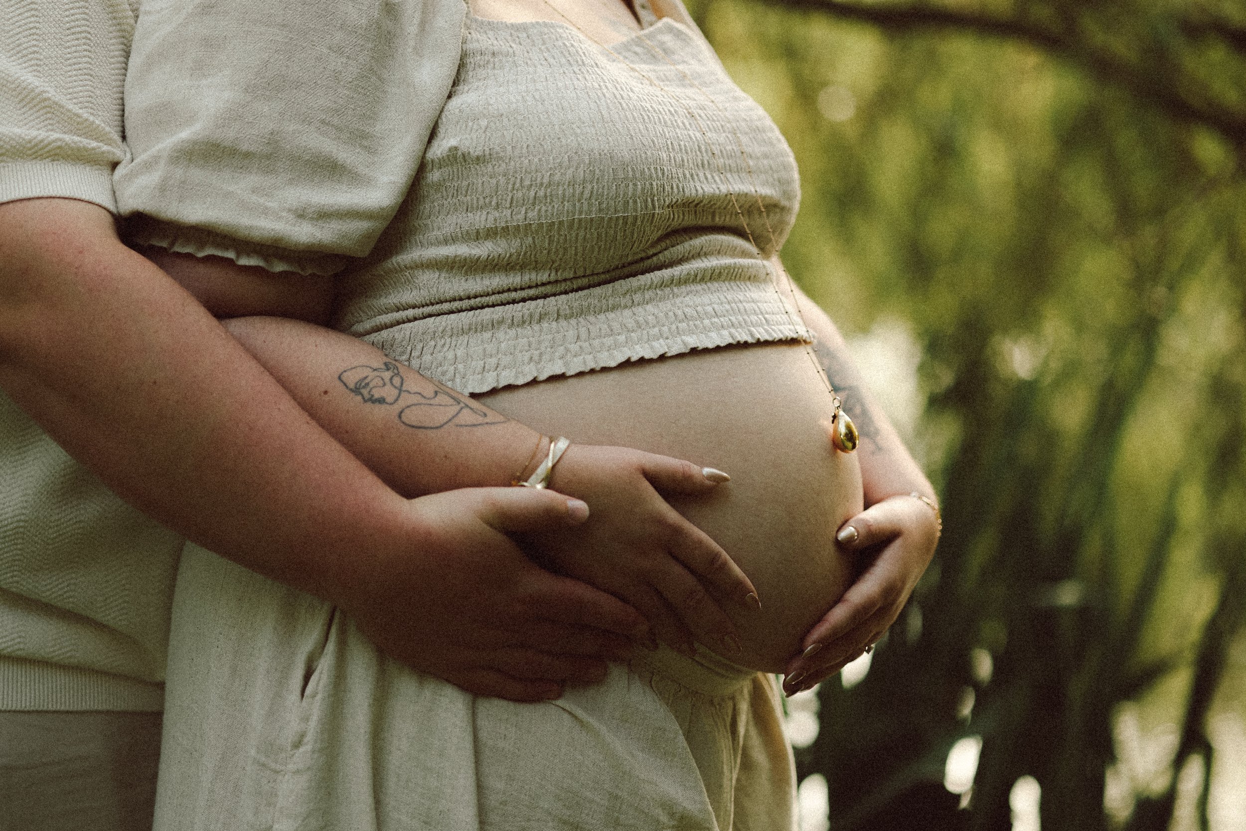 Une femme enceinte tenant son ventre avec les mains, entourée d'une autre personne dans un environnement extérieur avec des arbres.
