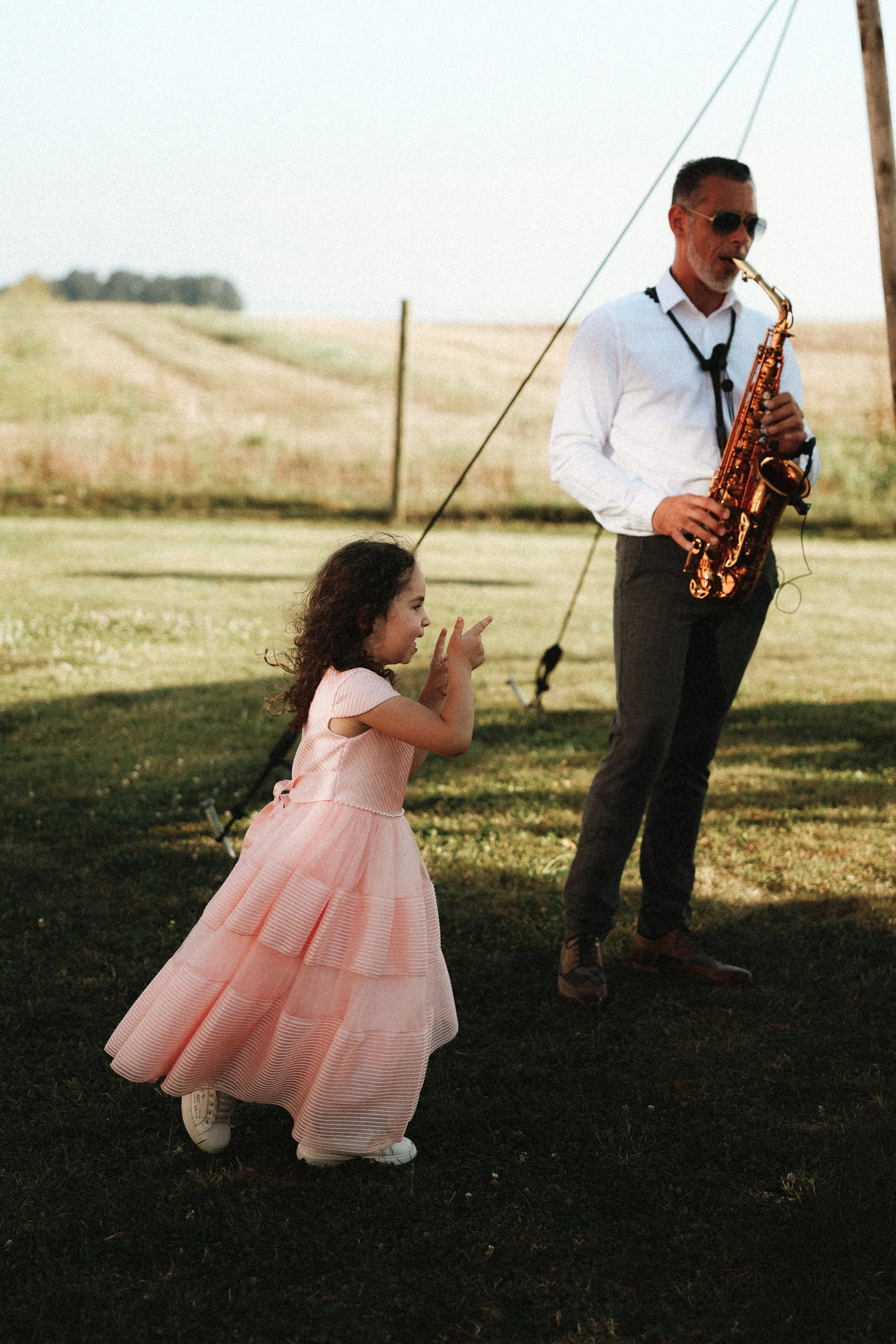 Une petite fille en robe rose danse devant un musicien jouant du saxophone dans un champ en plein air.
