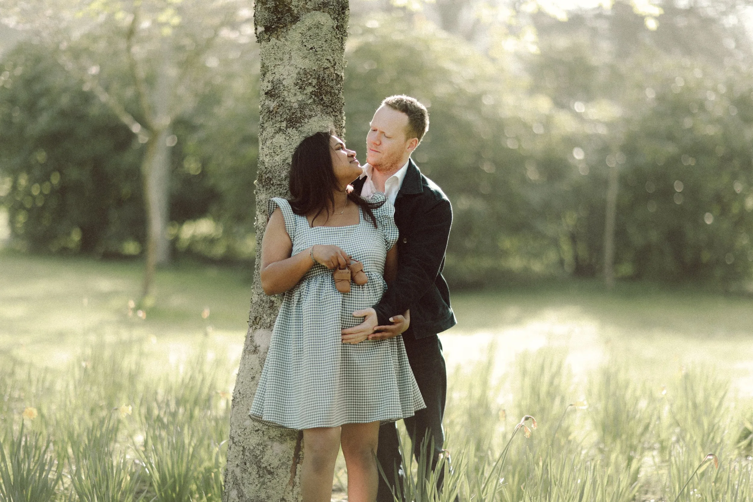 Un couple s'affiche tendrement dans un champ en extérieur, la femme enceinte se tient contre un arbre et l'homme la regarde avec amour.