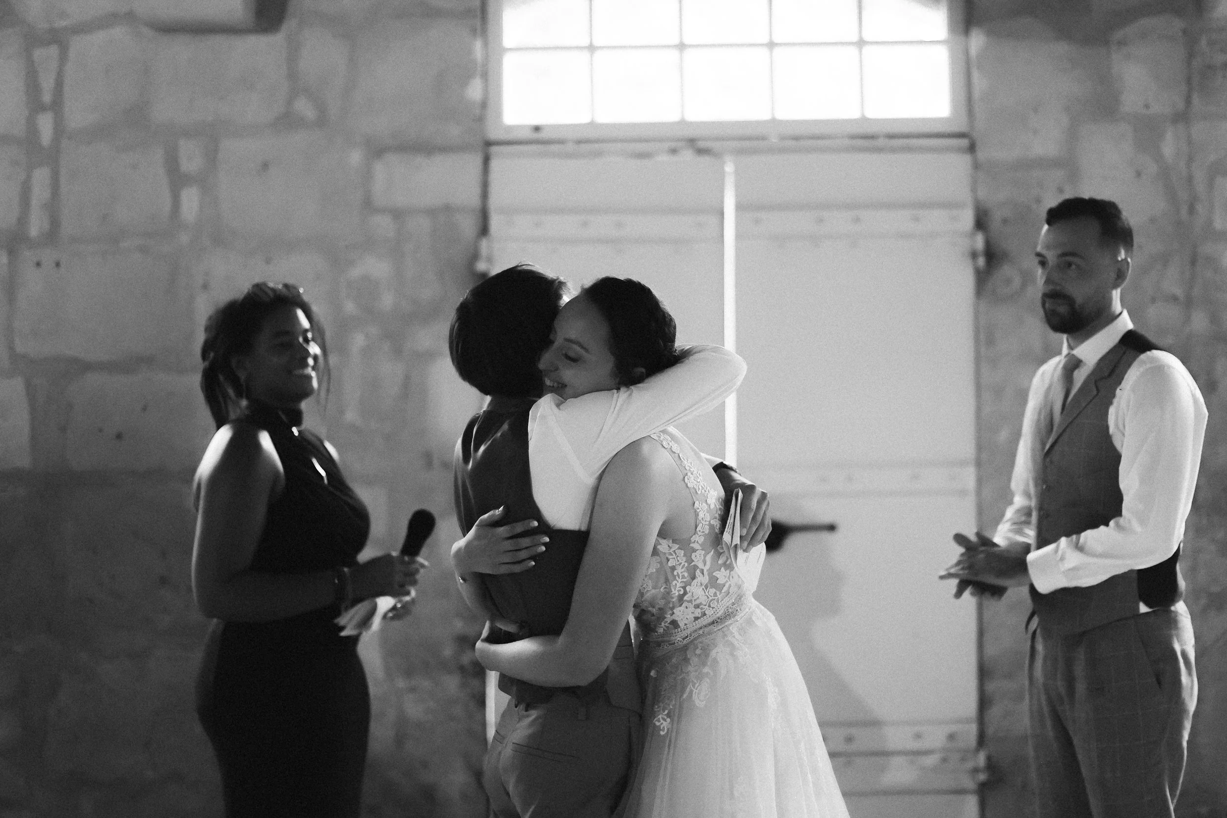 Deux femmes s'embrassent, l'une en robe de mariée, dans une salle avec murs en pierre, pendant qu'une autre femme souriante tient un micro et un homme observe. En arrière-plan, une grande porte en bois.