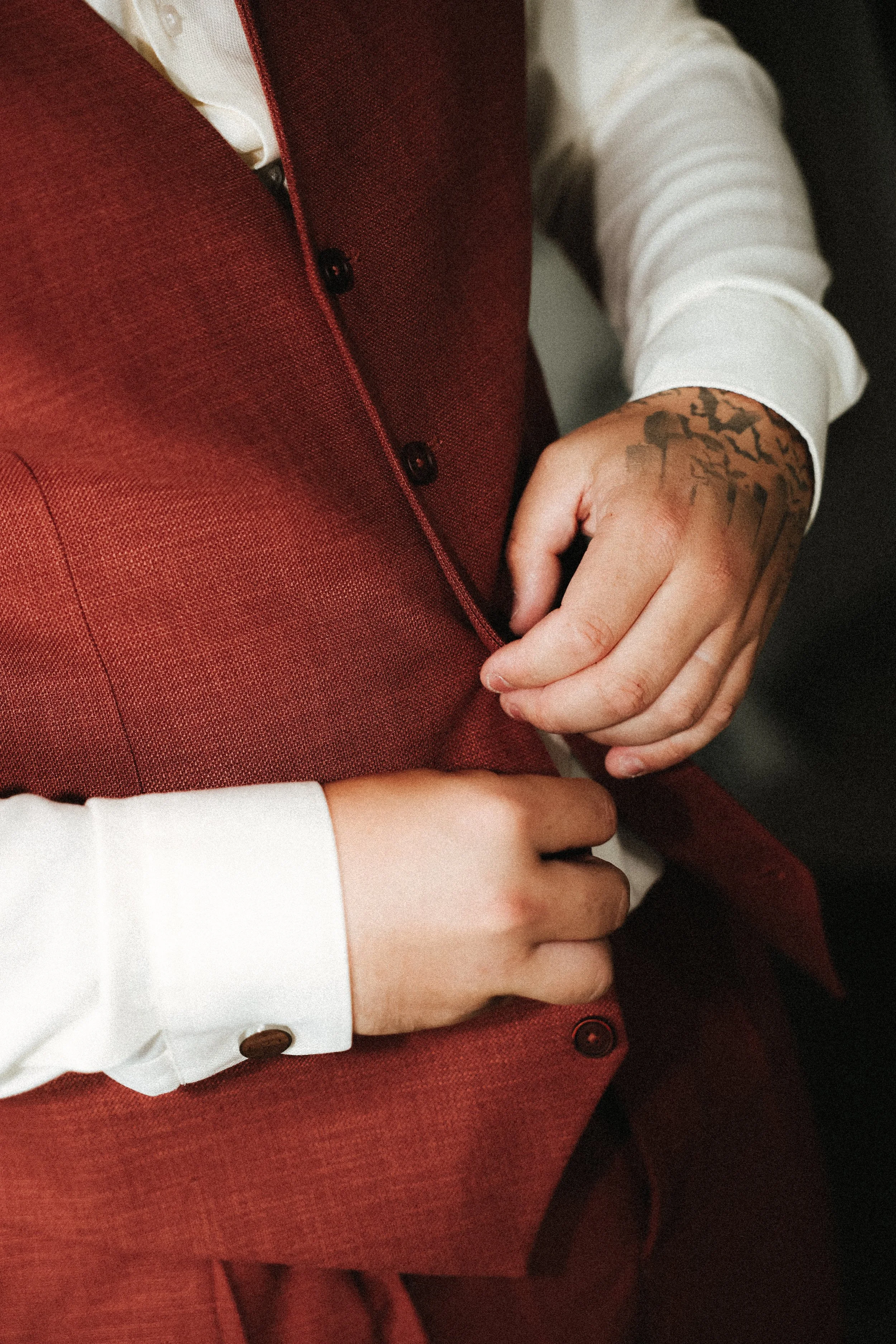 Gros plan sur un homme en costume rouge ajustant ses boutons, portant une chemise blanche et ayant un tatouage sur la main.