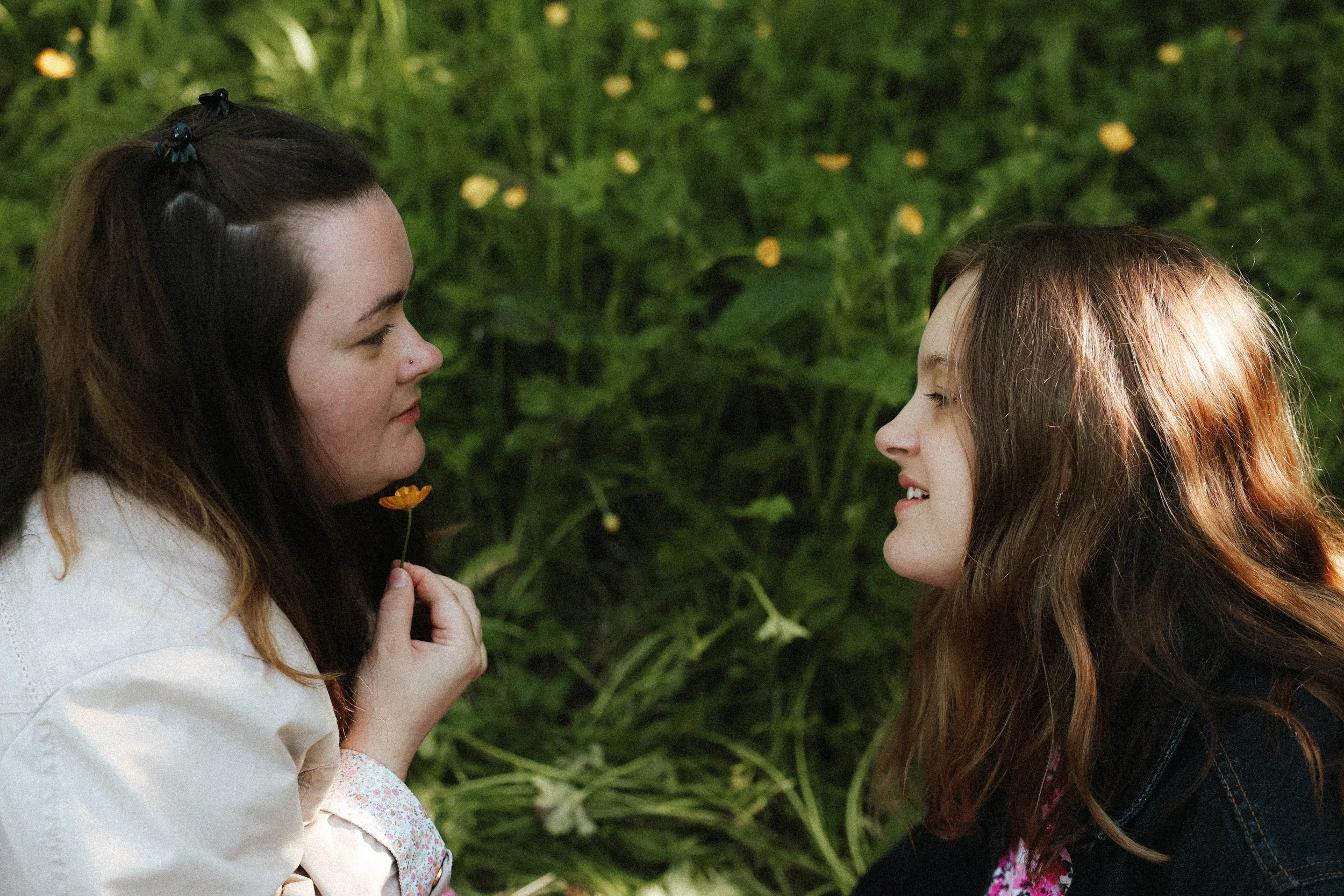 Deux jeunes filles regardent et sourient l'une à l'autre, avec un fond de végétation.