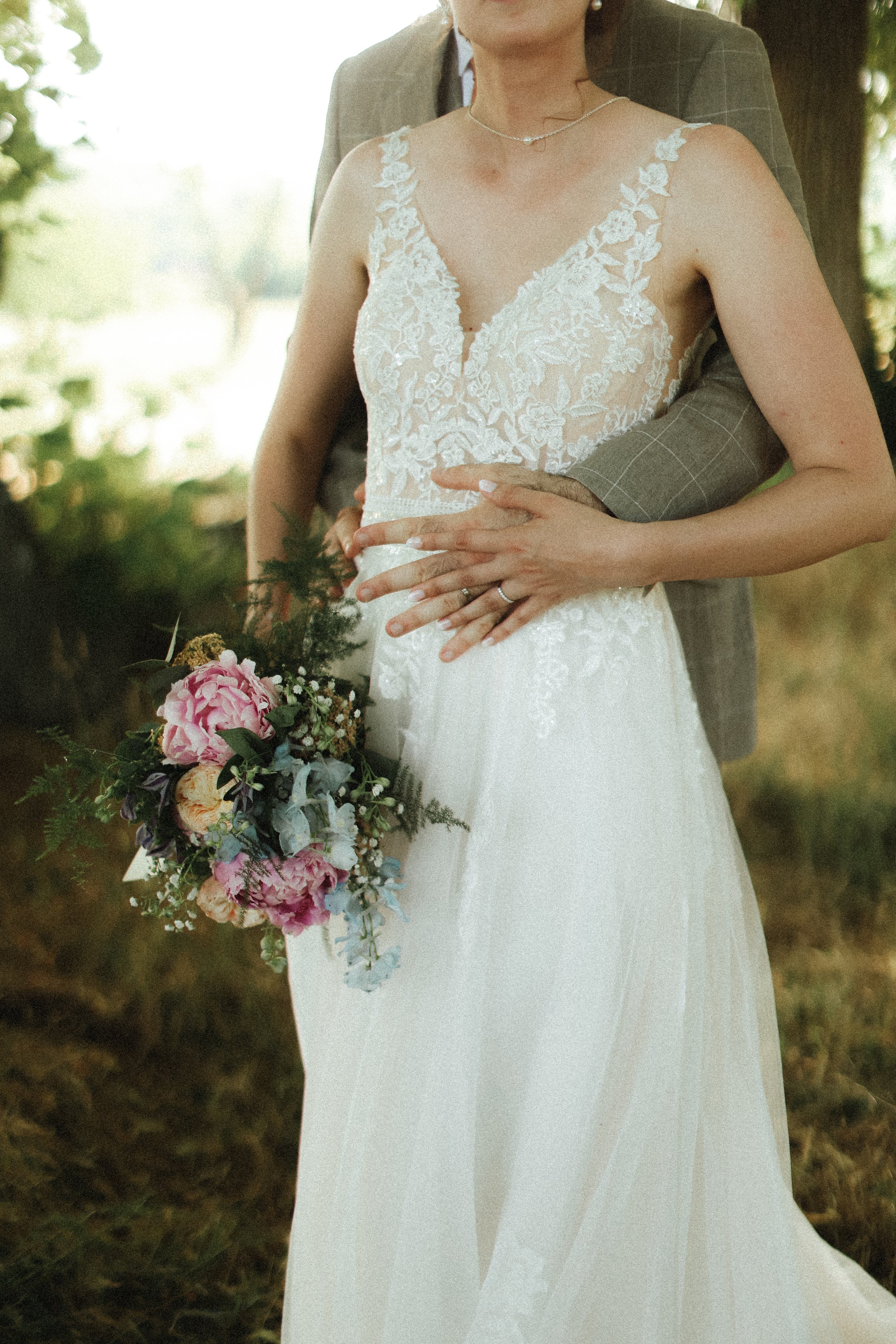 Un couple lors d'un mariage, la femme en robe de mariée blanche en dentelle, tenant un bouquet de fleurs colorées, l'homme en costume gris clair, entourés par un environnement verdoyant.