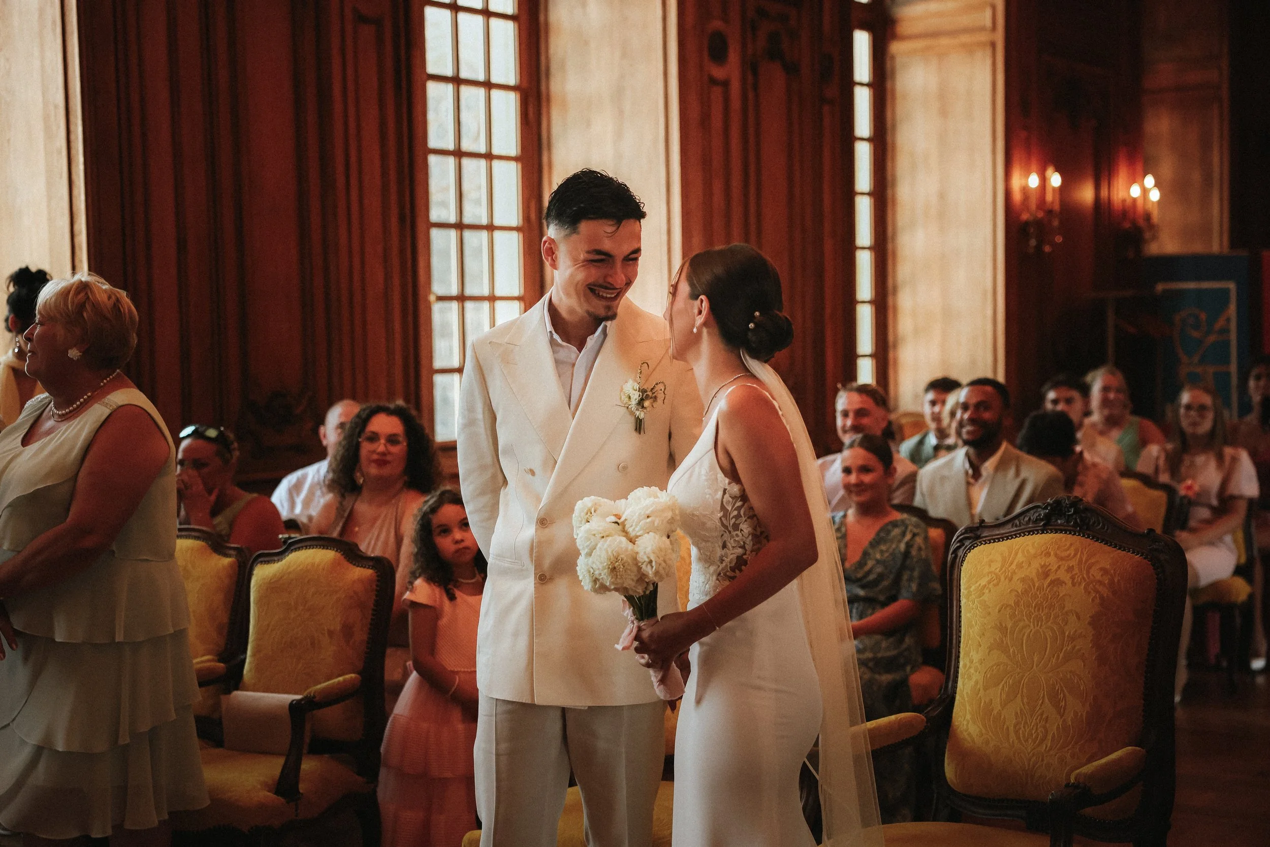 Un couple de mariés échangeant un regard dans une salle de cérémonie en bois, entouré de leurs invités qui les regardent avec émotion.