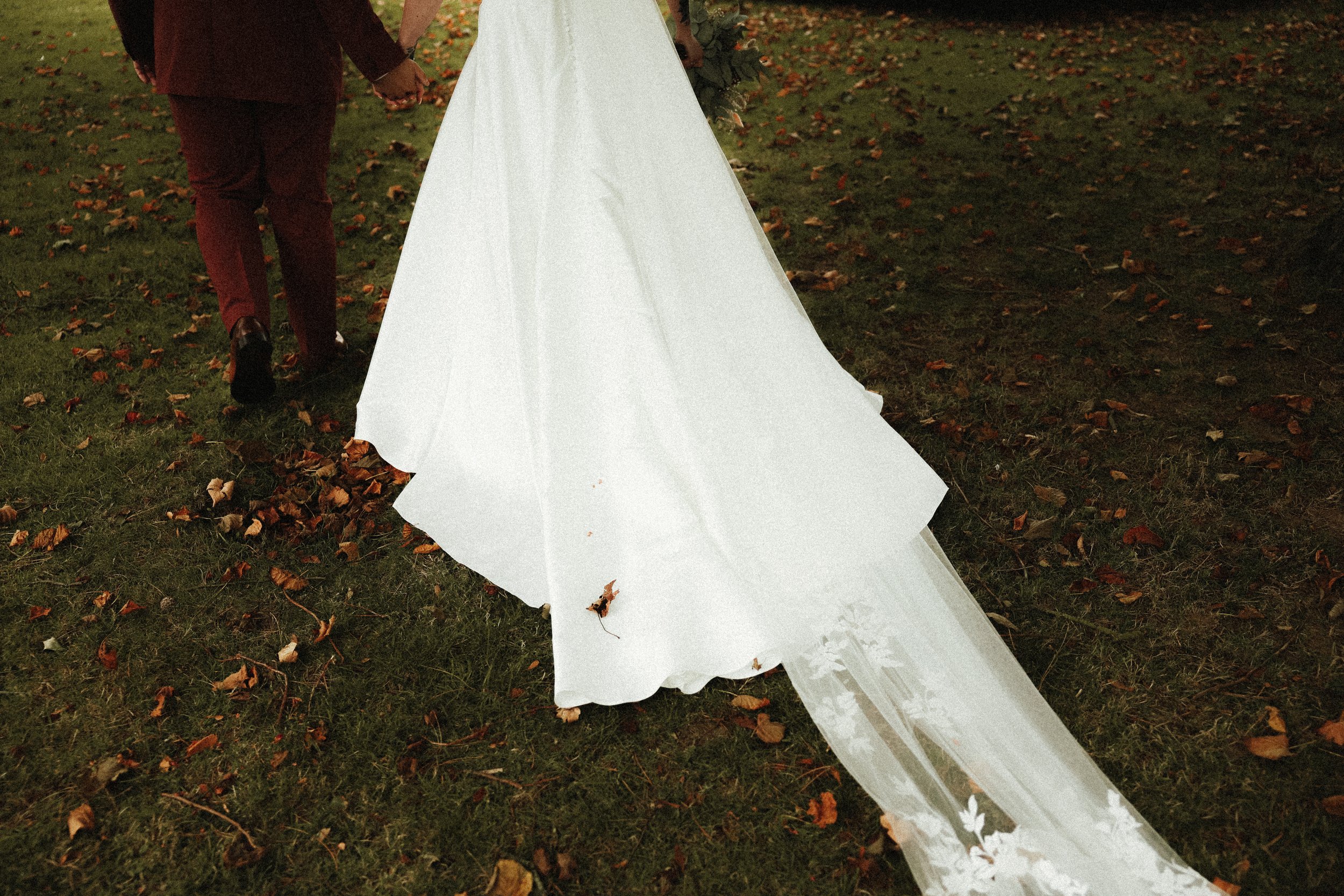 Un couple de mariés tient la main lors d'une cérémonie nuptiale en plein air, avec une robe de mariée blanche et un décor automnal avec des feuilles sur l'herbe.