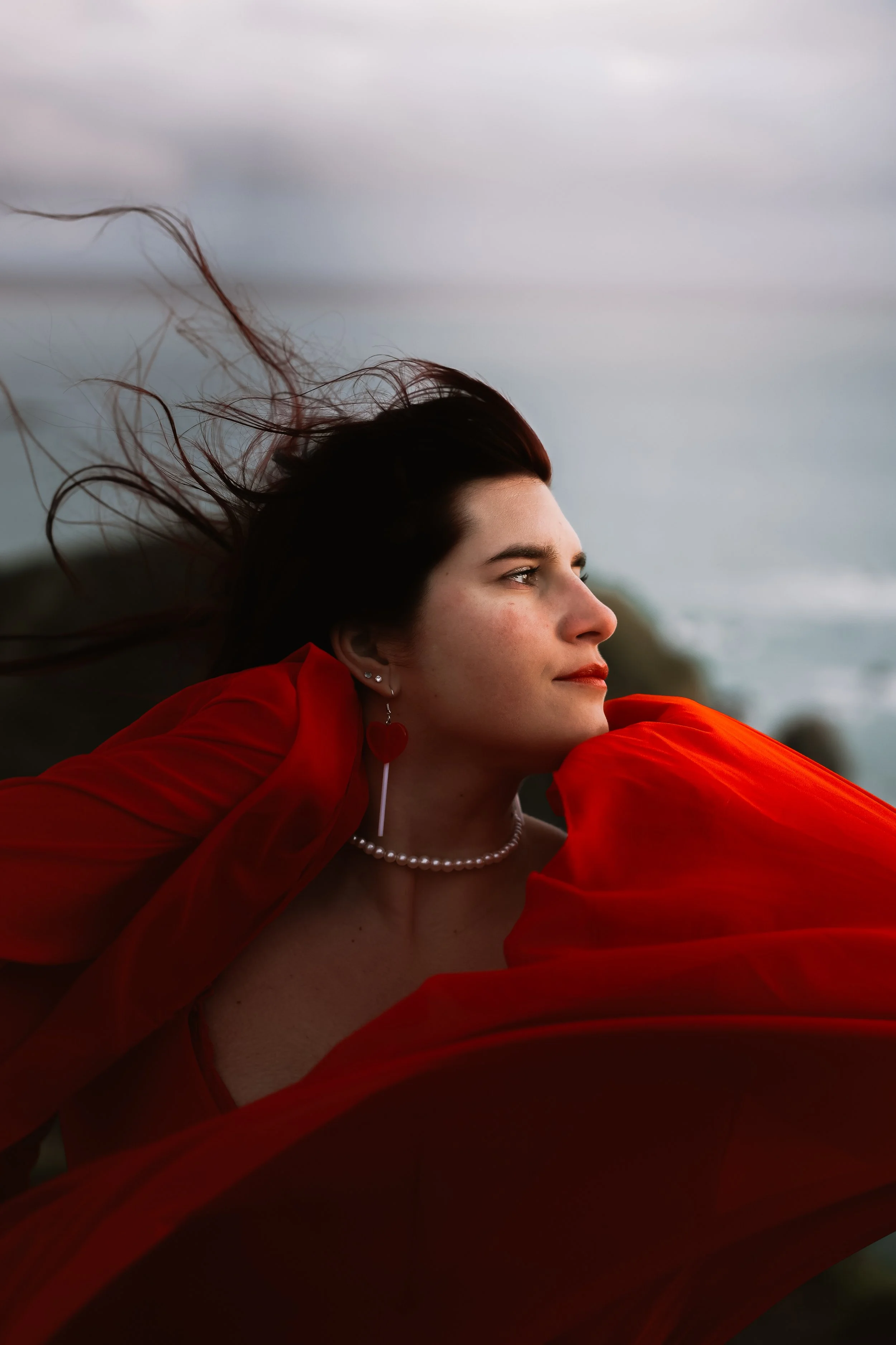 Une jeune femme avec des cheveux noirs, portant une robe rouge, des boucles d'oreilles en forme de cœur, une chaîne de perles, regarde vers la droite avec un ciel nuageux en arrière-plan.