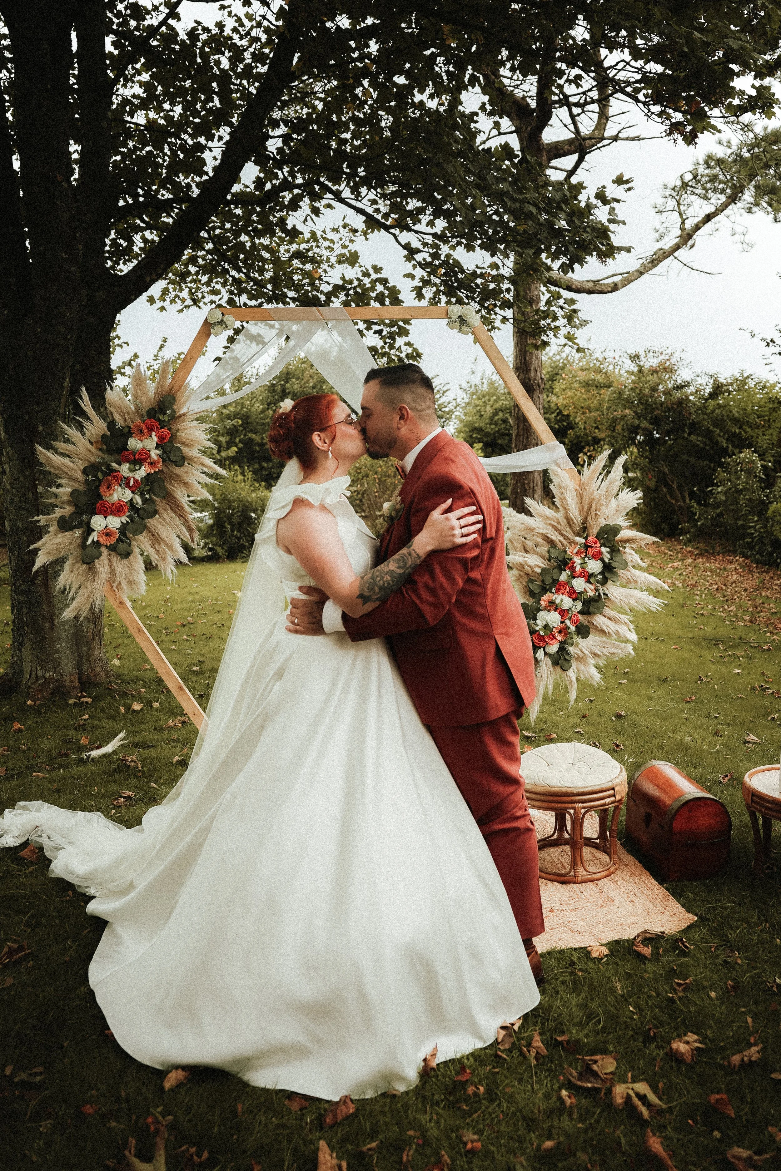 Un couple de mariés s'embrassant lors de leur mariage en plein air, avec un décor floral et un cadre en bois en forme d'hexagone.