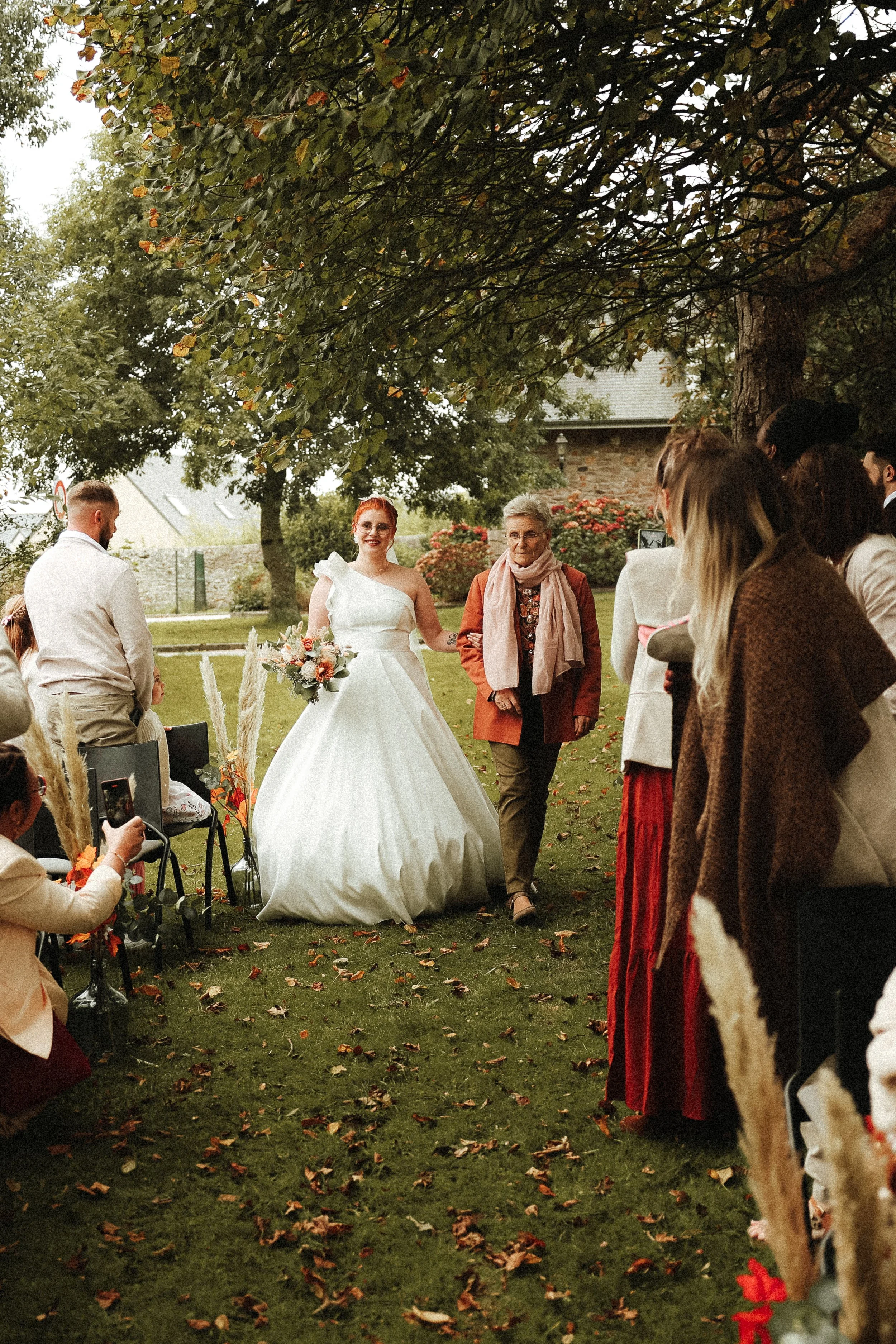 Une mariée en robe blanche souriante, portant un bouquet, marche avec une femme âgée lors d'un mariage en plein air sous un arbre, entourée de invités.
