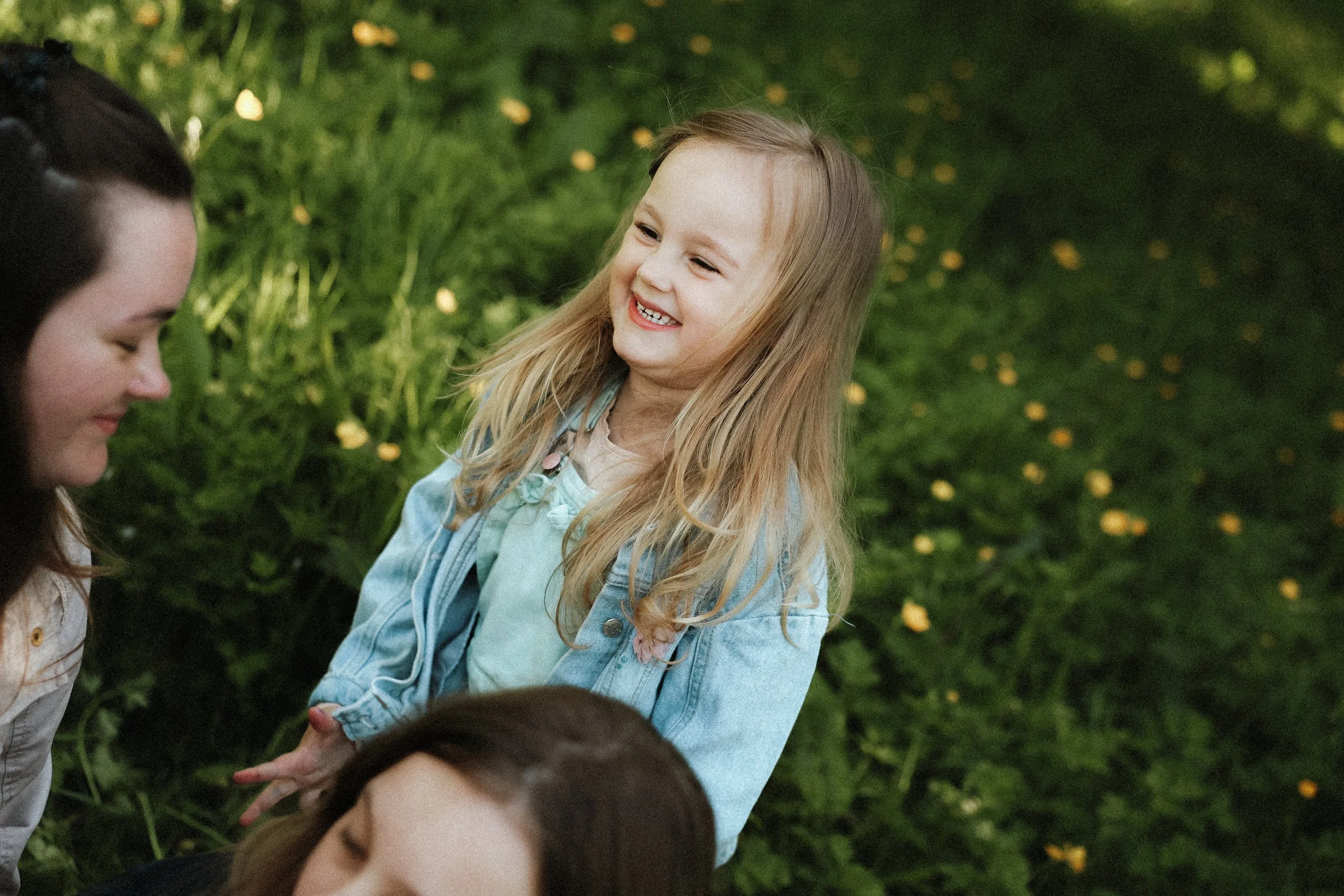 Une jeune fille aux longs cheveux blonds sourit dans un environnement naturel avec des buissons remplis de petites fleurs jaunes.