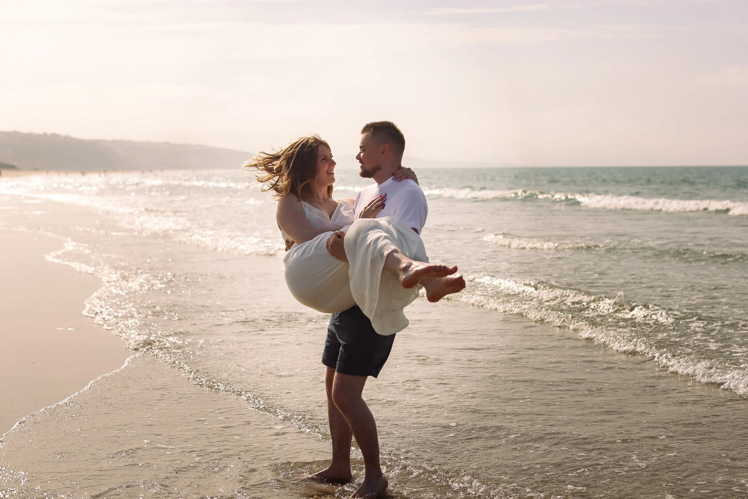 Un homme porte une femme dans ses bras sur la plage, ils sourient et se regardent, avec la mer et le ciel en arrière-plan.