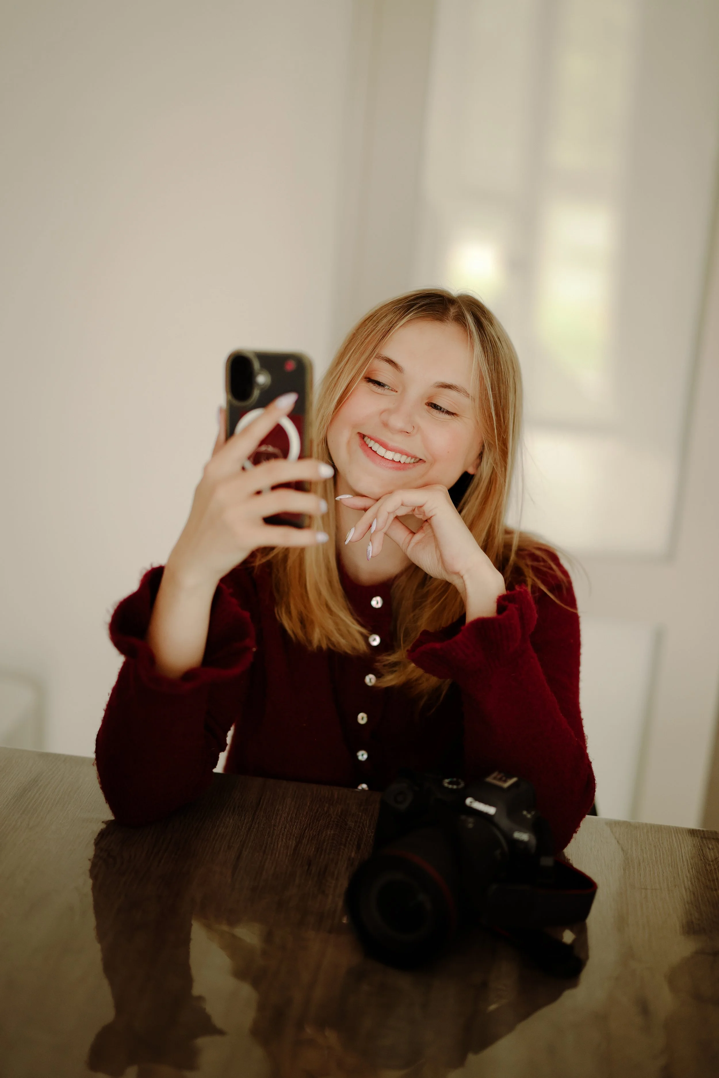 Jeune femme rieuse avec long cheveux blonds, portant un pull rouge à boutons, se prenant en selfie avec un téléphone, caméra sur la table en face d'elle.