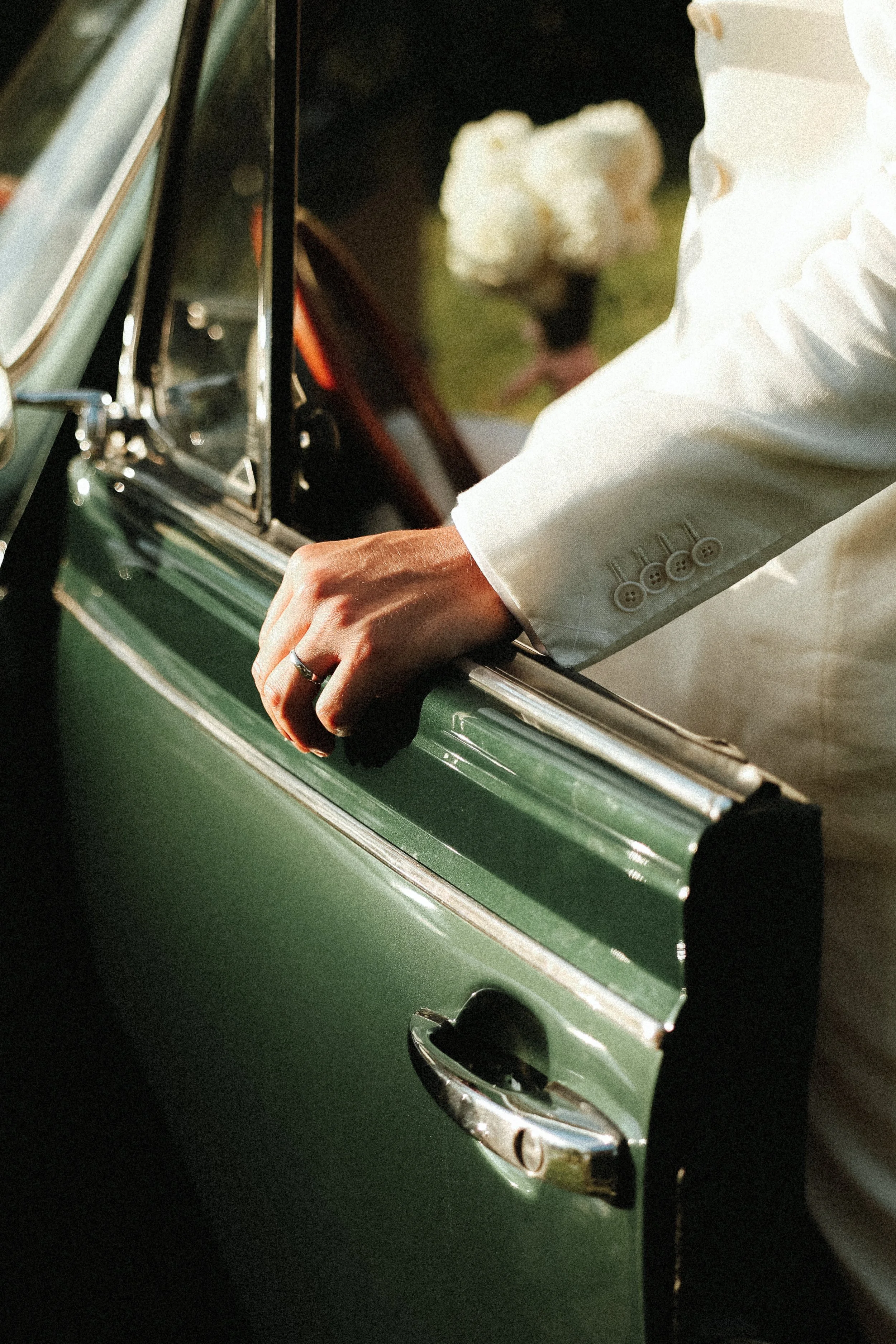 Main d'une personne en costume blanc sur la portière d'une voiture verte, avec une bouquet de fleurs en arrière-plan.