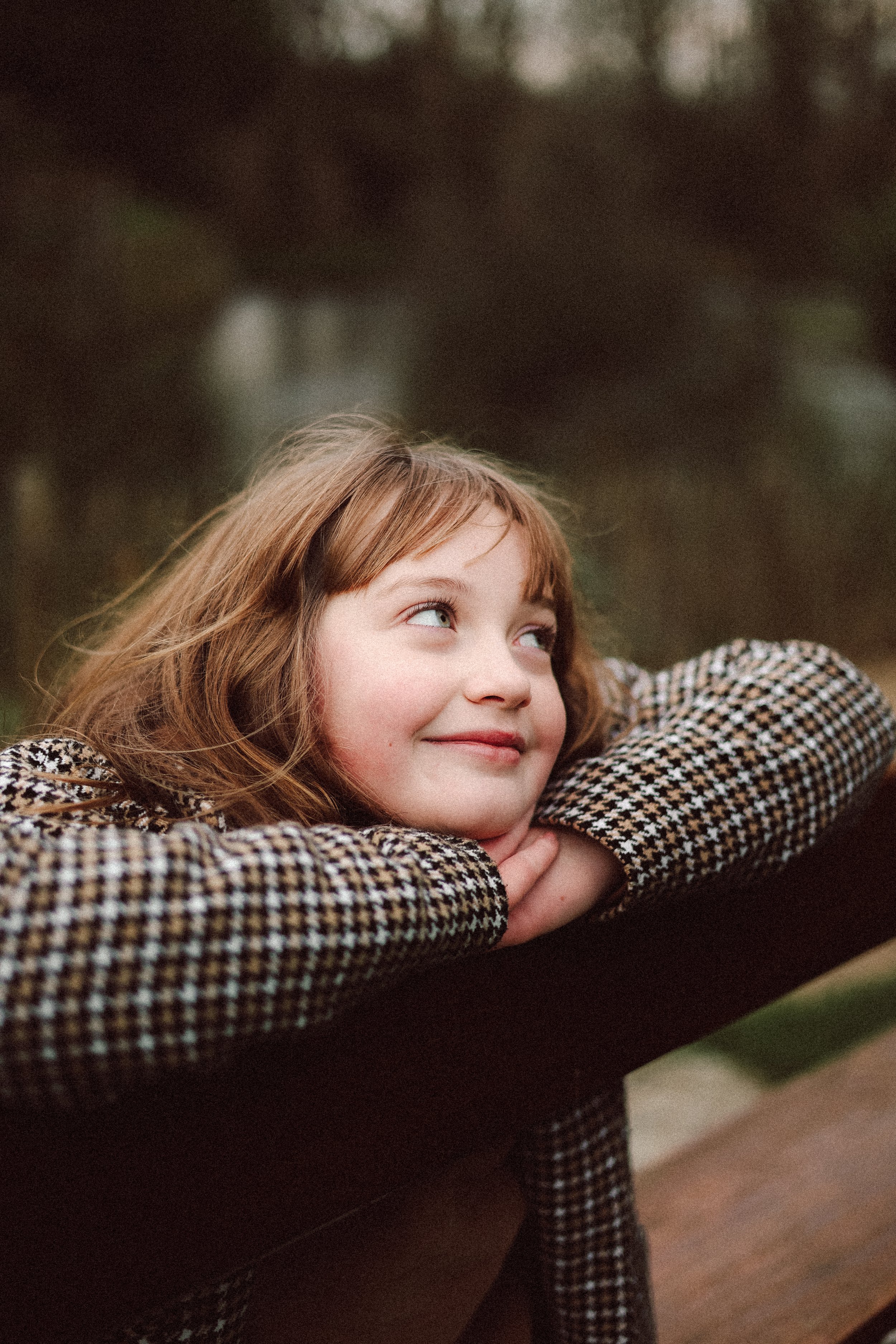 Jeune fille aux cheveux bruns avec des yeux bleus, portant un pull à carreaux, repose sa tête sur ses bras croisés sur un banc en bois, regardant vers le haut avec un sourire dans un environnement extérieur.