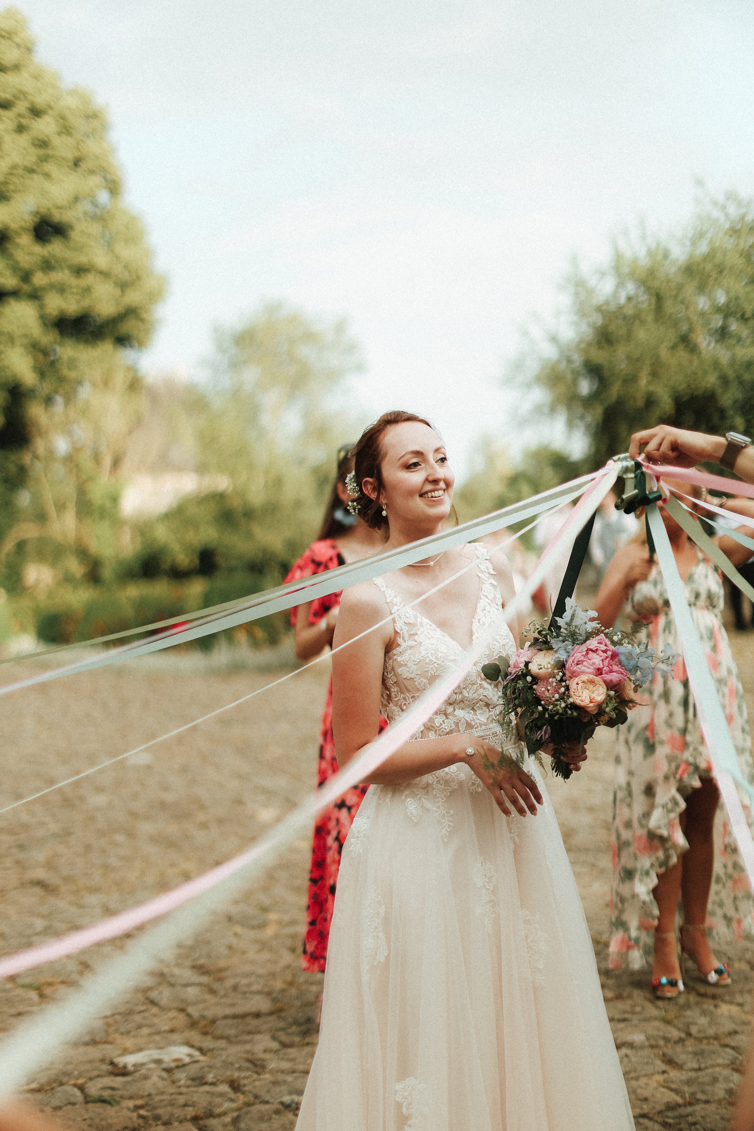 Une mariée souriante portant une robe de mariage blanche, tenant un bouquet de fleurs, lors d'une fête de mariage en plein air avec des invités et des décorations de rubans colorés.