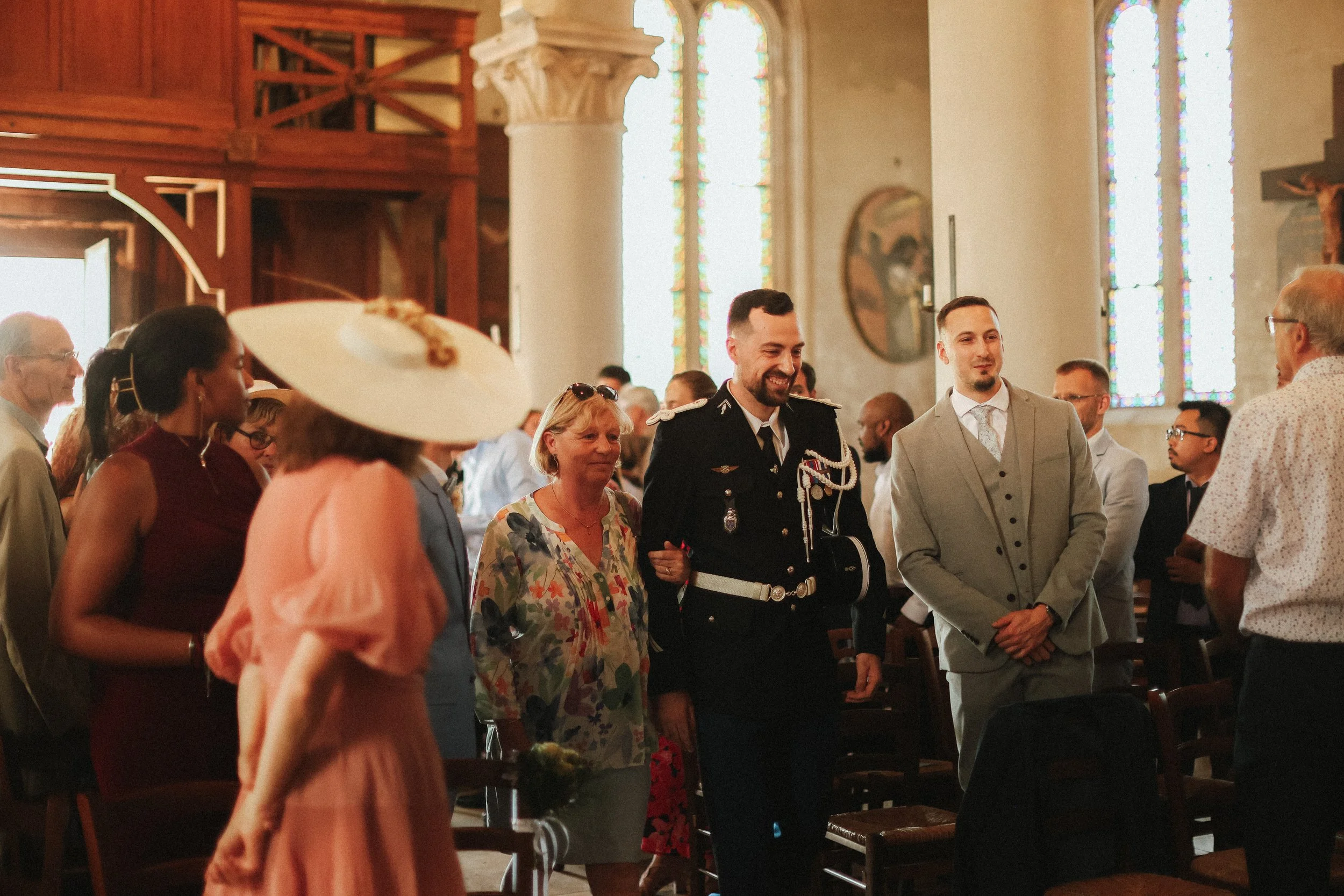 Des personnes en pleine célébration dans une chapelle, avec un homme en uniforme militaire au centre, entouré de personnes habillées de façon formelle et élégante, lors d'une cérémonie ou mariage.