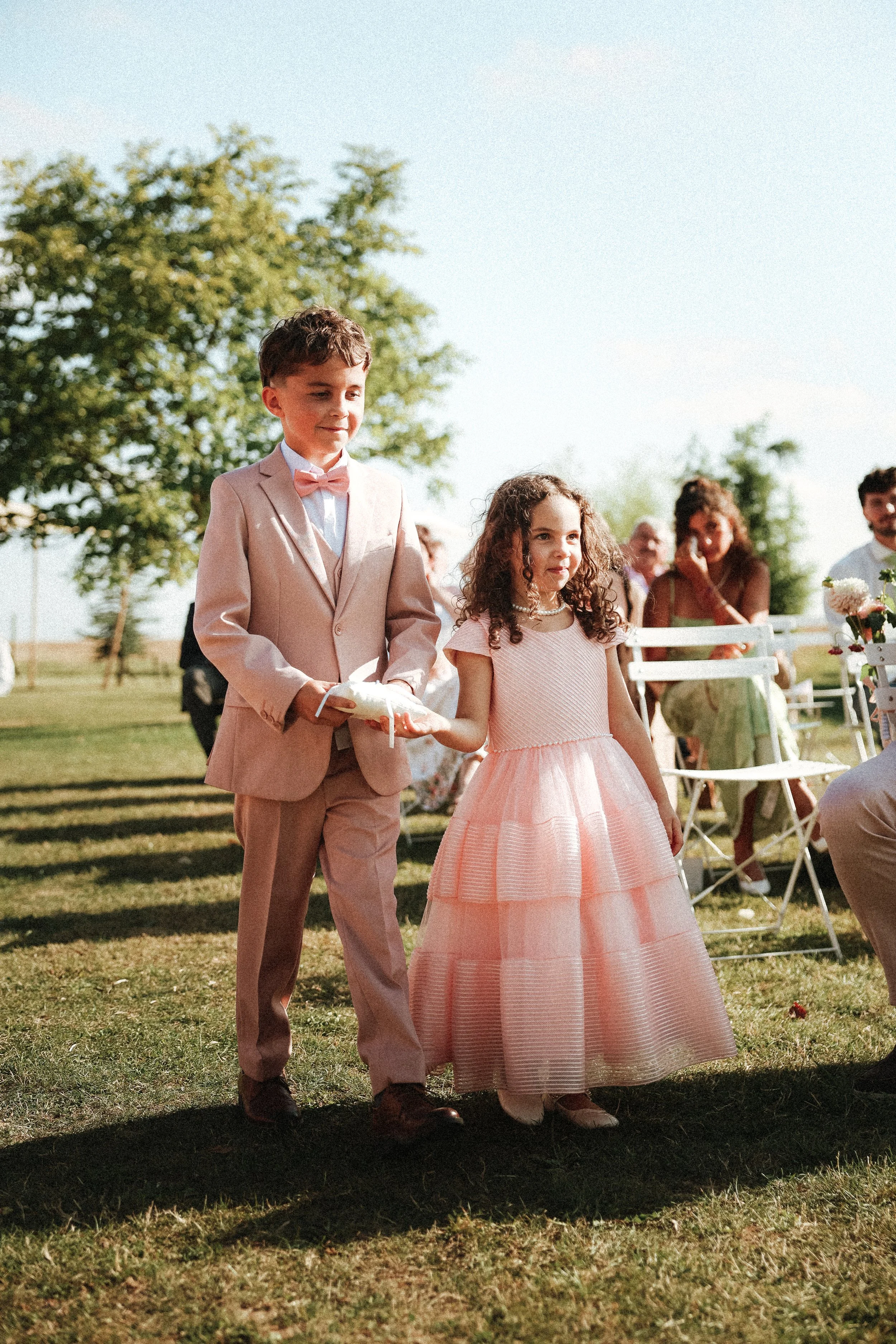 Deux enfants en vêtements formels, un garçon en costume beige avec un nœud papillon rose, une fille en robe rose, marchent main dans la main lors d'une cérémonie extérieure en plein-air, avec des invités assis à côté.