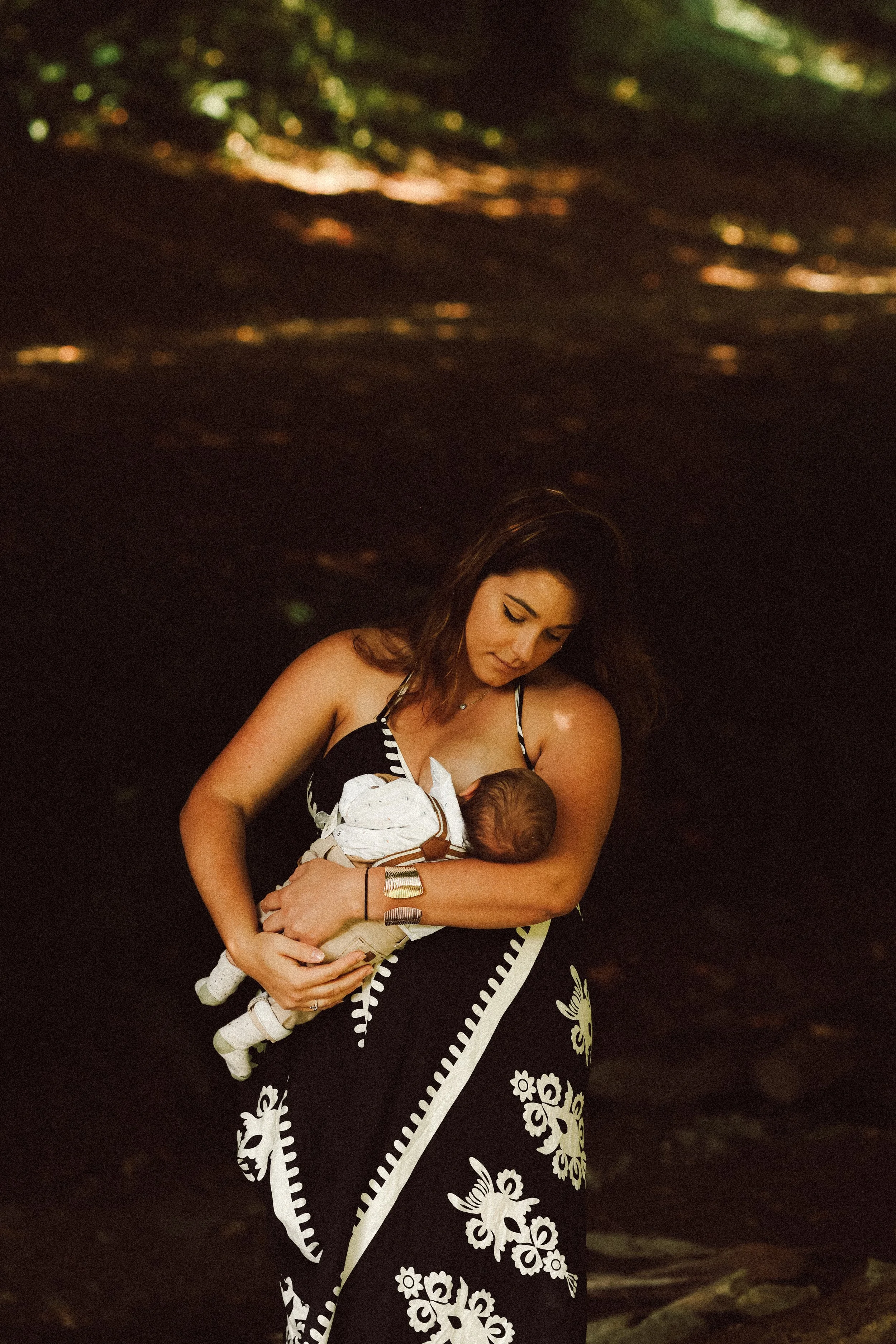 Femme tenant un bébé dans ses bras, vêillée d'une robe noire avec des motifs blancs, dans un environnement sombre.