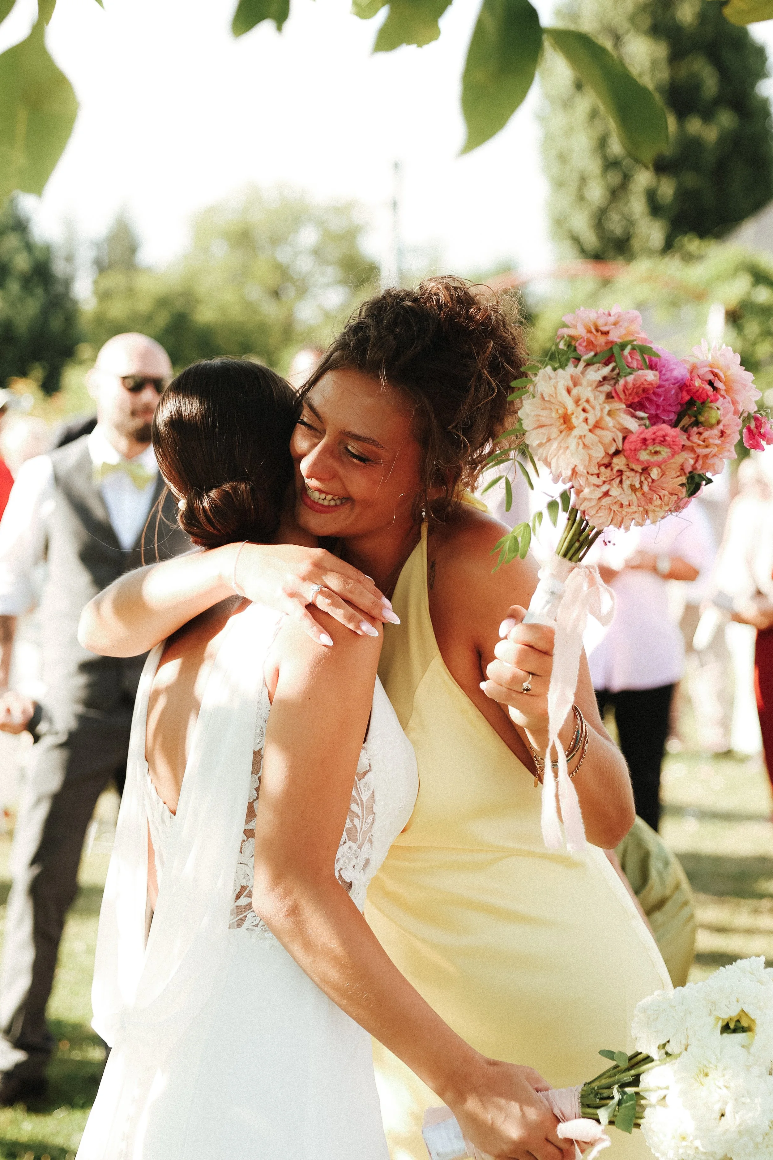 Deux femmes s'embrassent lors d'un mariage, l'une tenant un bouquet de fleurs roses et l'autre portant une robe blanche, dans un jardin en plein air.