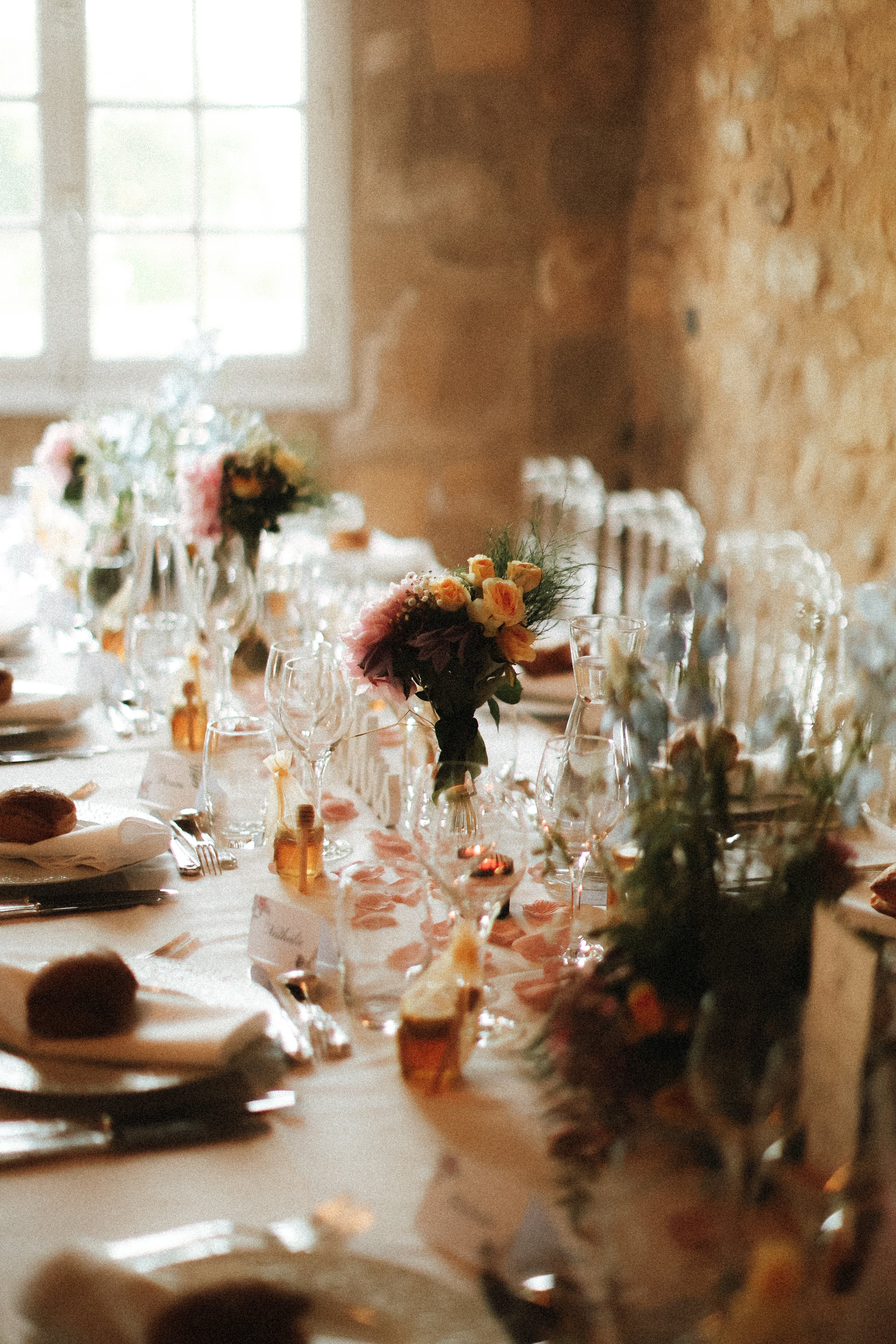 Table de fête décorée avec des fleurs et des verres en cristal, dans une salle avec pierre apparente et fenêtre lumineuse.