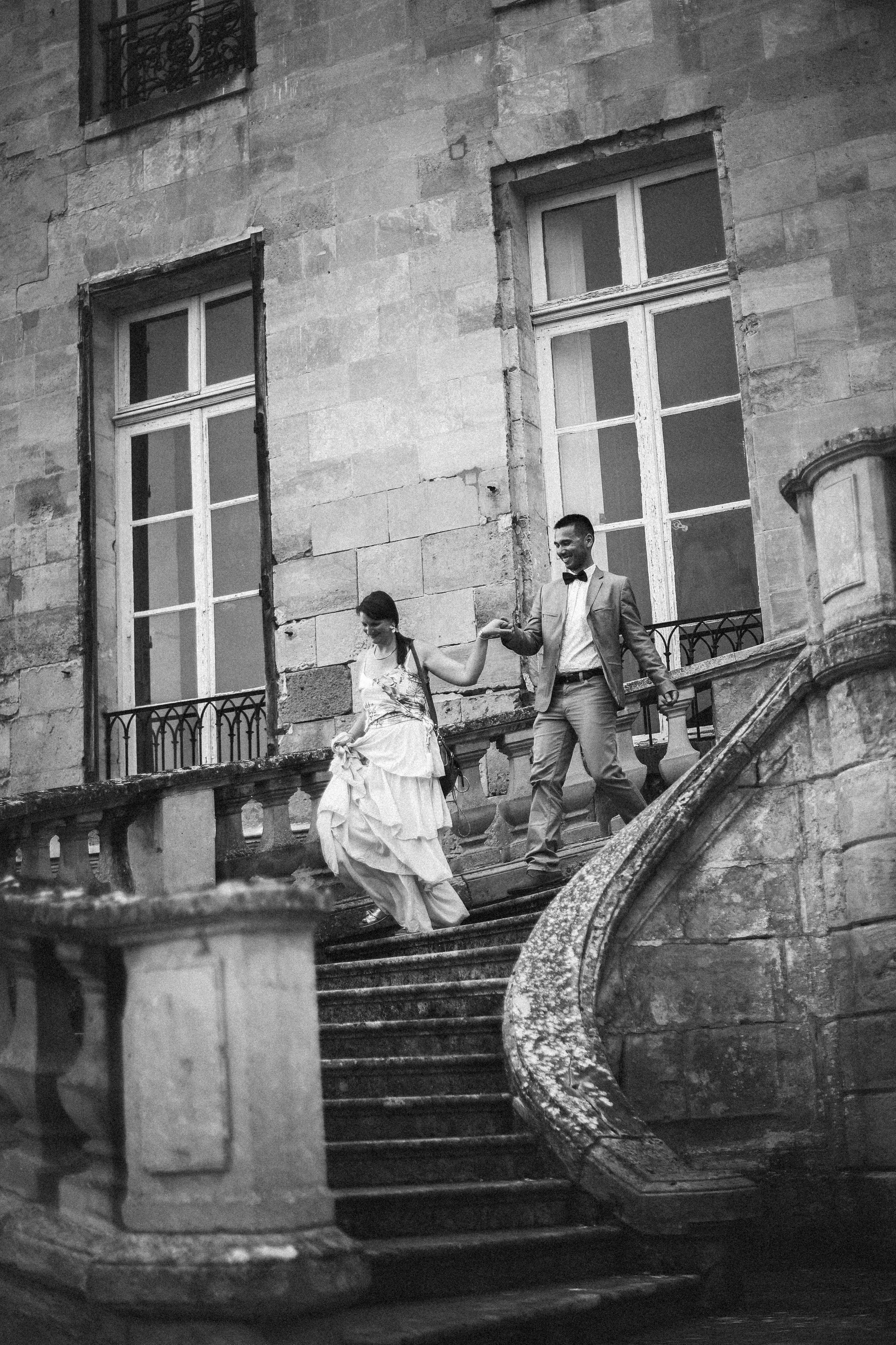 Un couple en habits de mariage descend un escalier en pierre devant un bâtiment ancien en pierre. La femme porte une robe de mariée blanche, et l'homme porte un costume, tous deux souriants.