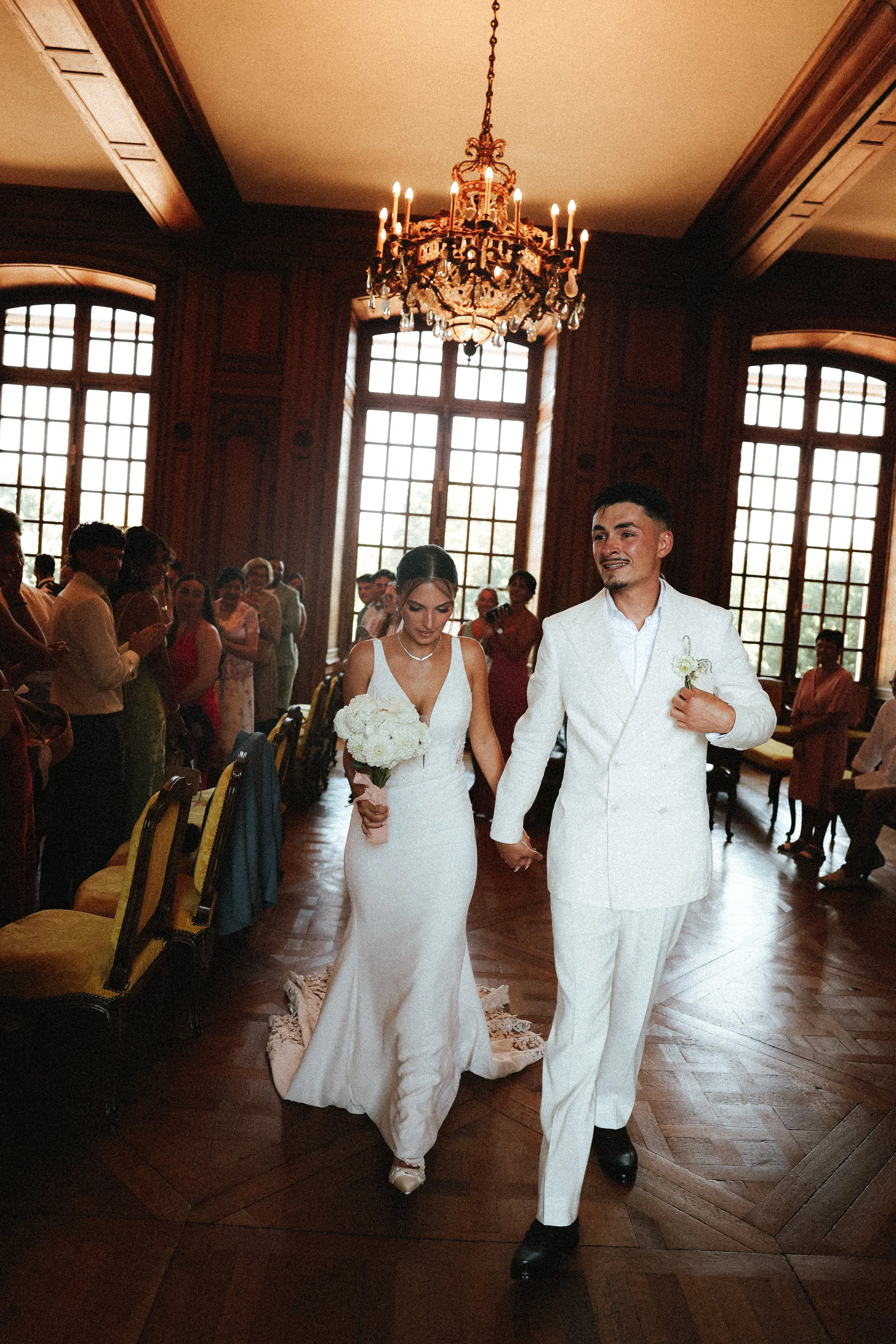 Un couple de mariés marche dans une salle de cérémonie élégante avec des invités en arrière-plan, intérieur en bois avec grandes fenêtres et un chandelier au plafond.