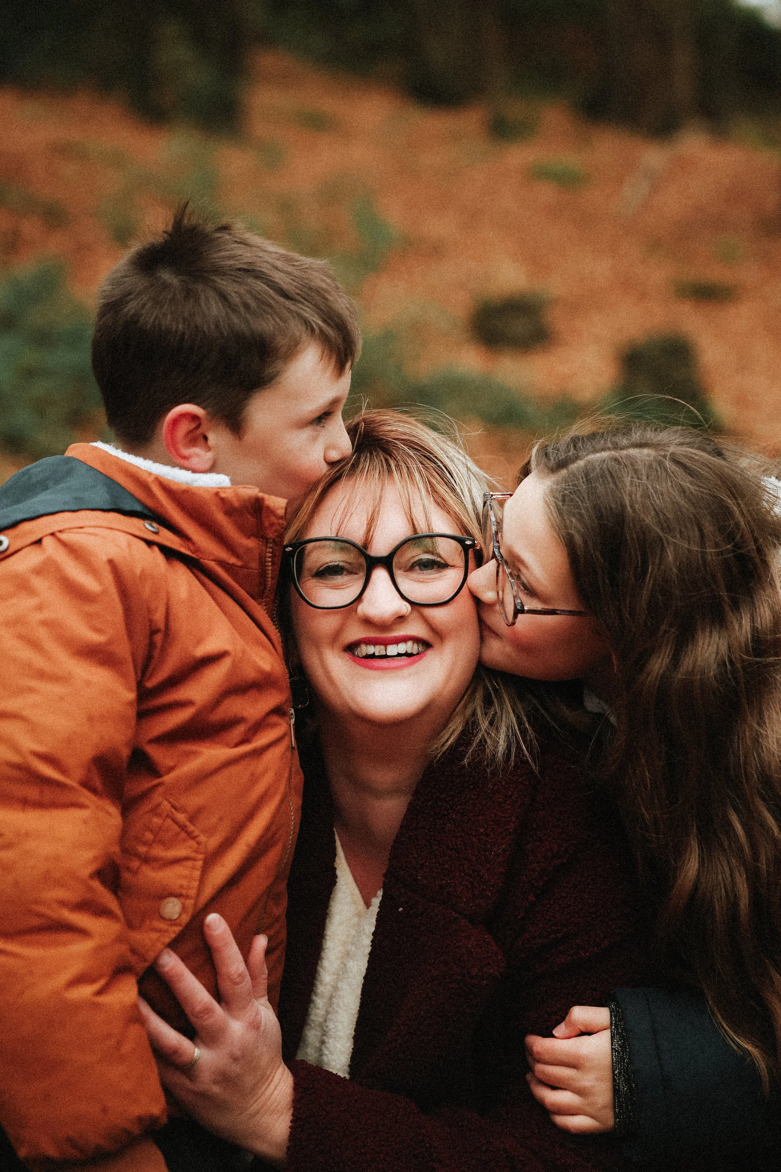 Une femme porte des lunettes noires et sourit tandis qu'un garçon et une autre femme l'embrassent sur les joues dans une scène chaleureuse en plein air.