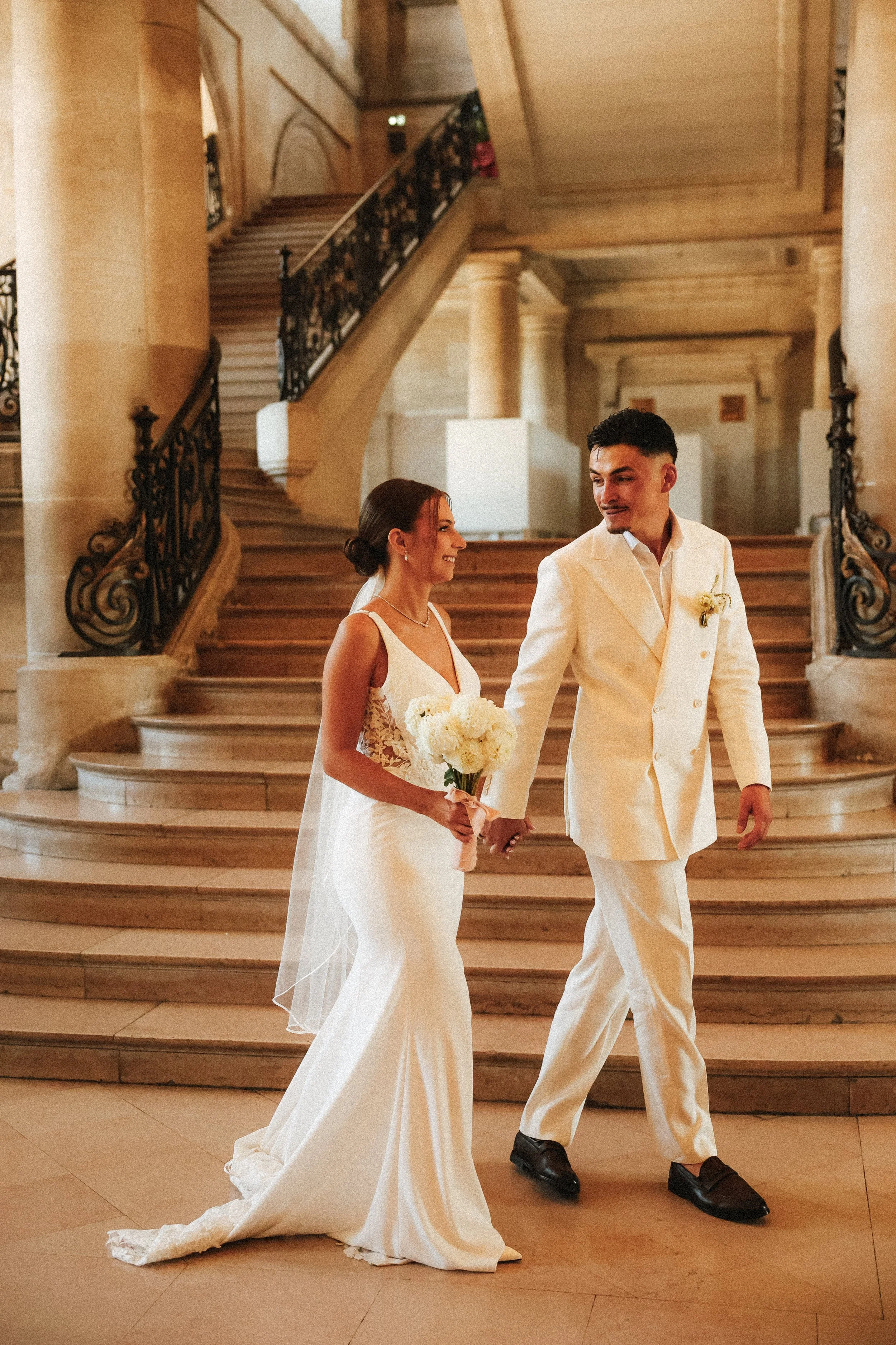 Un couple de mariés marche dans un hall élégant, la femme portant une robe de mariée blanche et tenant un bouquet de fleurs, l'homme en costume blanc. Ils sourient et se tiennent par la main.