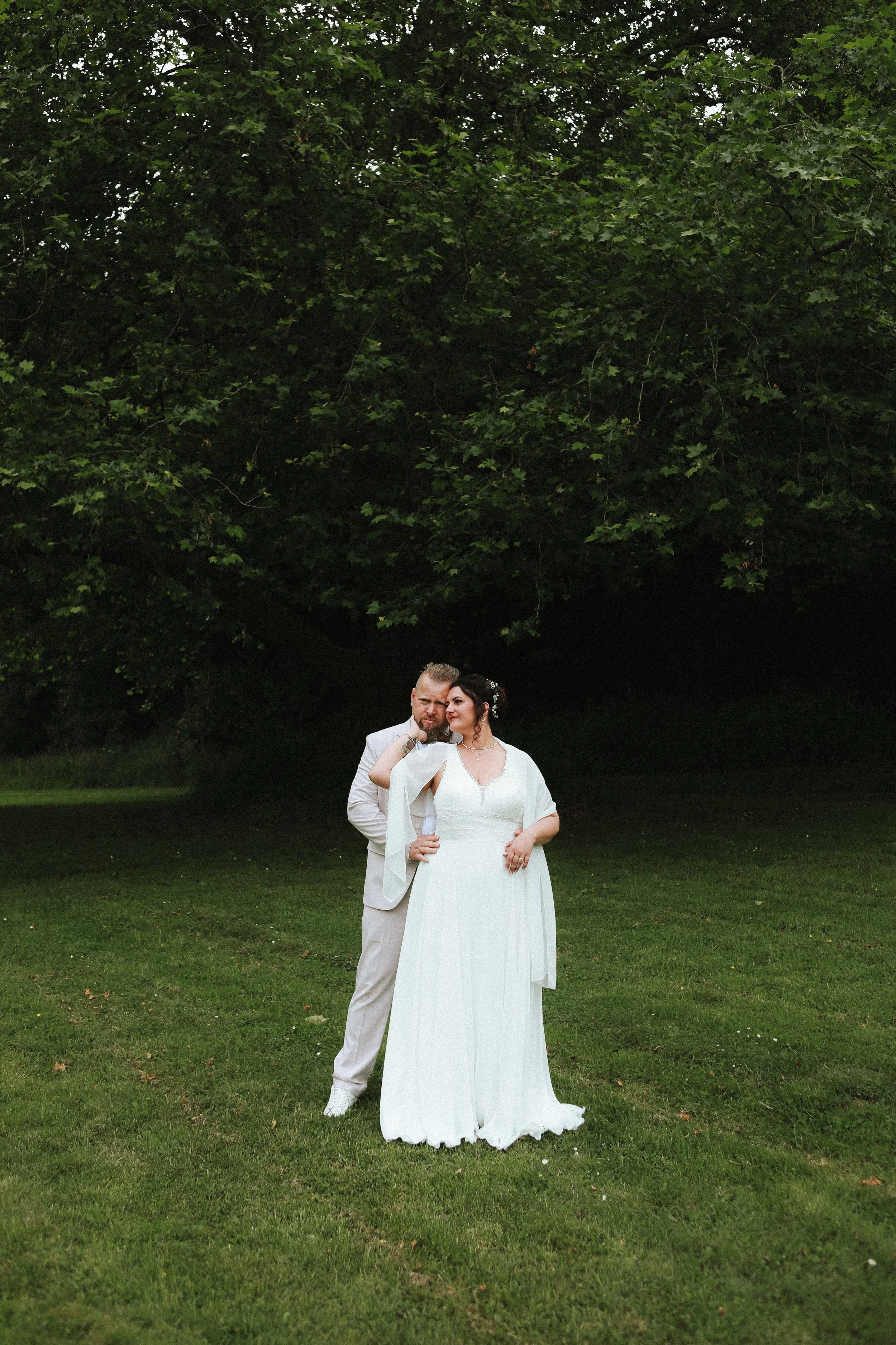Un couple en tenue de mariage posant dans un parc verdoyant sous un arbre volumineux.