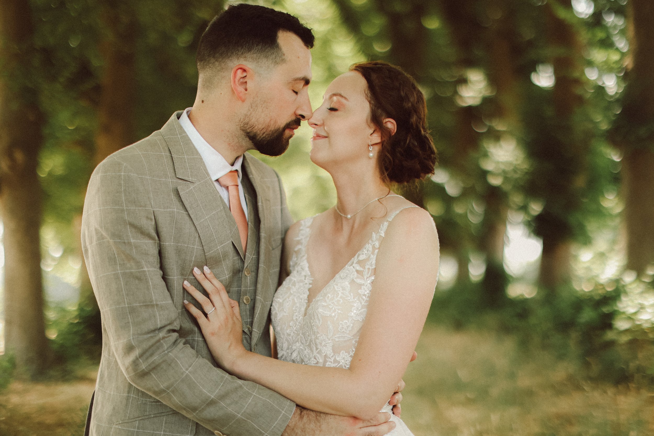 Un couple de mariés s'embrassant tendrement en forêt, homme en costume gris à carreaux et femme en robe de mariage blanche.