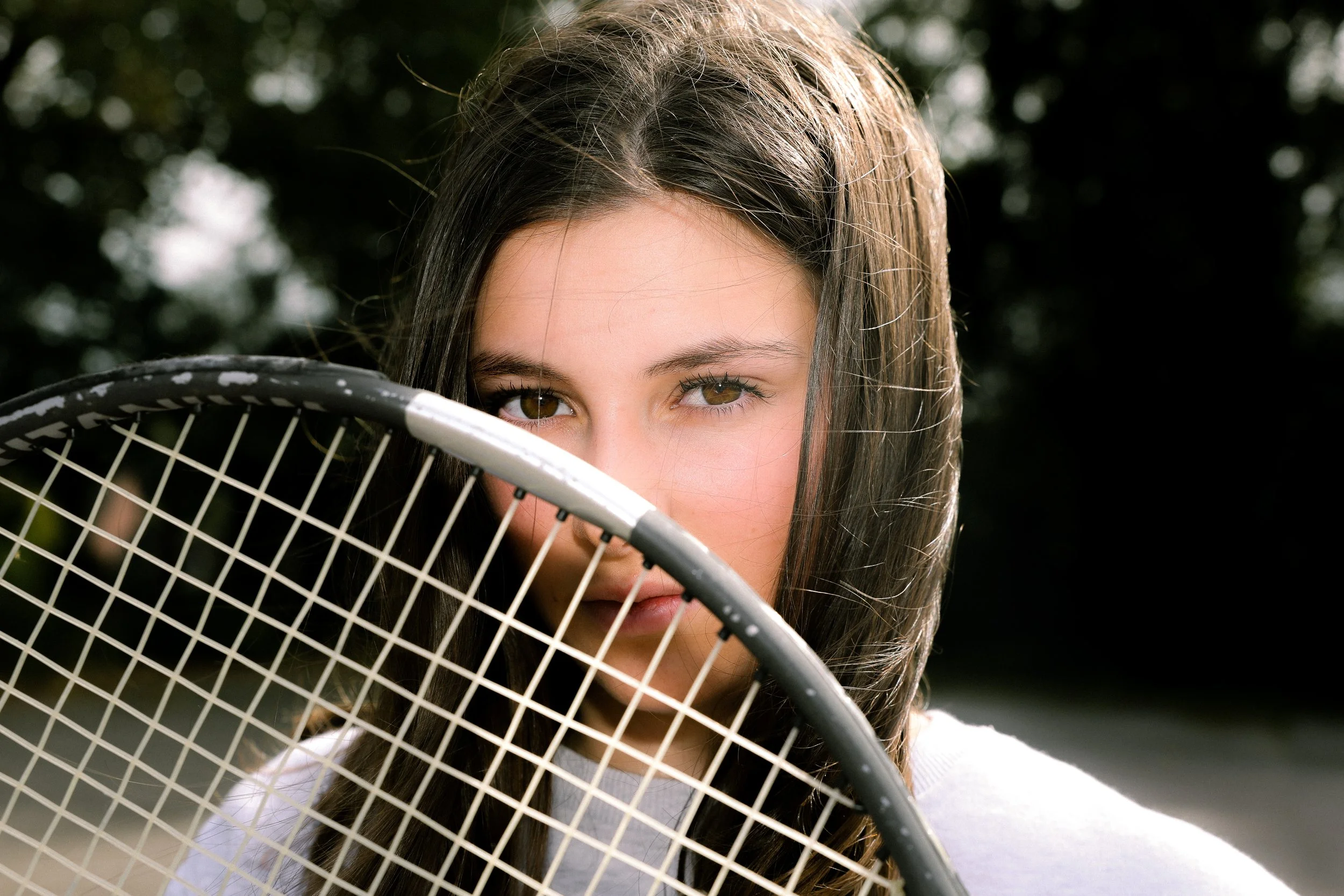 Une jeune fille aux yeux marron, tenant une raquette de tennis, se tenant à l'extérieur avec un fond verdoyant.